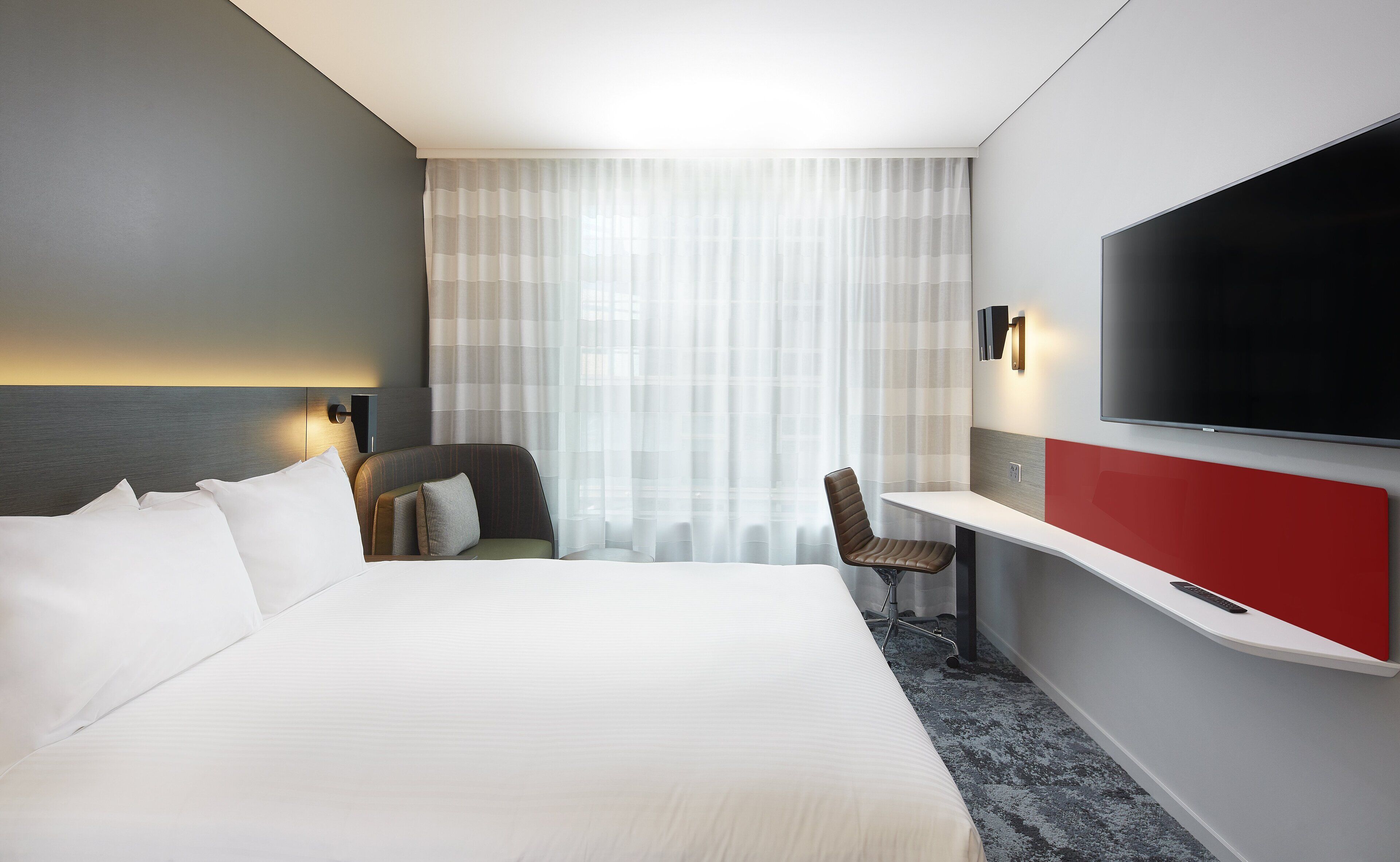 A hotel room with a large bed, a desk, a chair, a TV, and a window with sheer curtains.