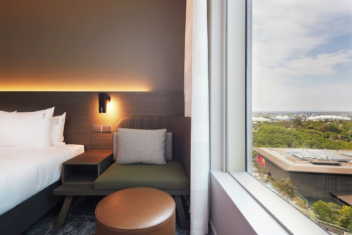 Hotellihuone, jossa on sänky, oleskelualue tyynyineen ja pieni pöytä sekä ikkunasta avautuva näkymä kaupunkimaisemaan.
