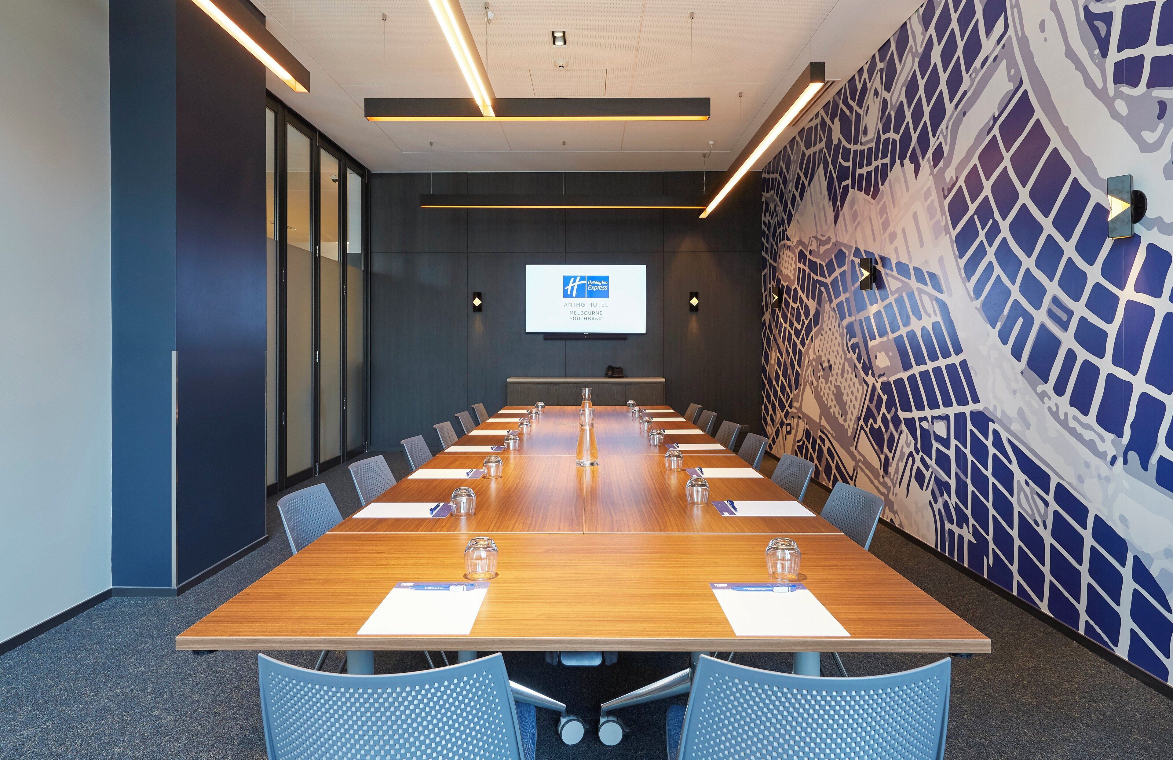 A conference room with a long table, chairs, a projector screen, and a patterned wall.