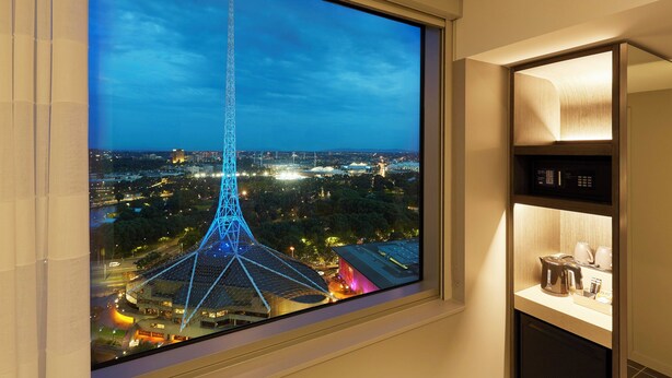 A room with a large window overlooking a cityscape at night, featuring a prominent tower and a well-lit interior with a coffee maker and a shelf. - Holiday Inn Express Melbourne Southbank by IHG (Southbank)