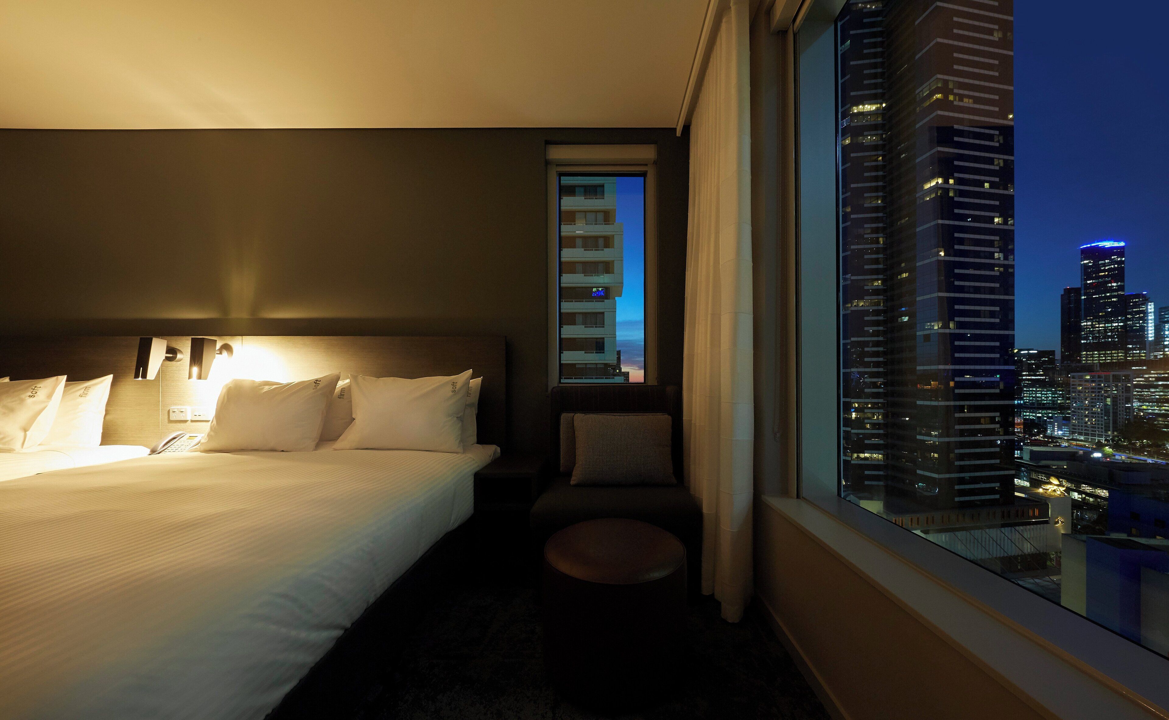 A hotel room with a bed, a bedside table, a small stool, and a window with a cityscape view.