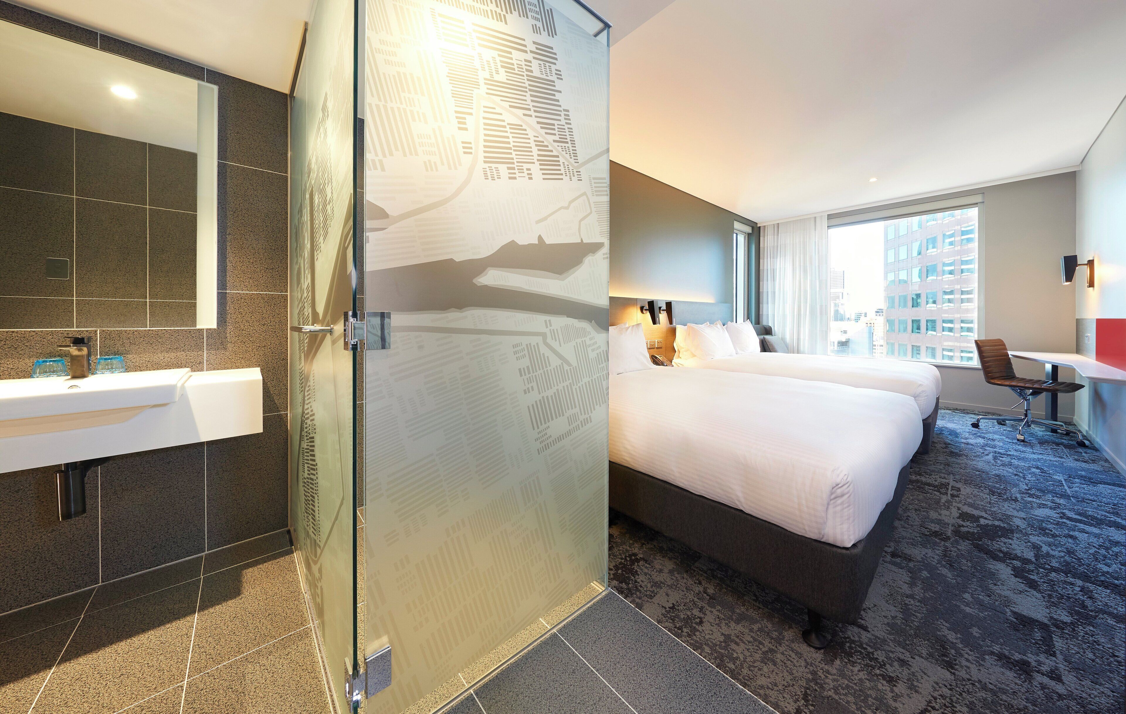 Une chambre d’hôtel dotée d’une douche en verre, d’un lit, d’une table de chevet, d’une chaise et d’une grande fenêtre donnant sur la ville.