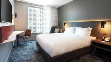 Ein Hotelzimmer mit einem großen Bett, einem Schreibtisch, einem Sessel, einem Fernseher und einem Fenster mit Blick auf Gebäude.