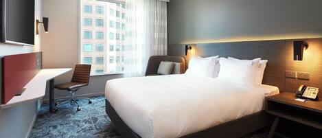 A hotel room with a large bed, a desk, a chair, a TV, and a window with a view of buildings.