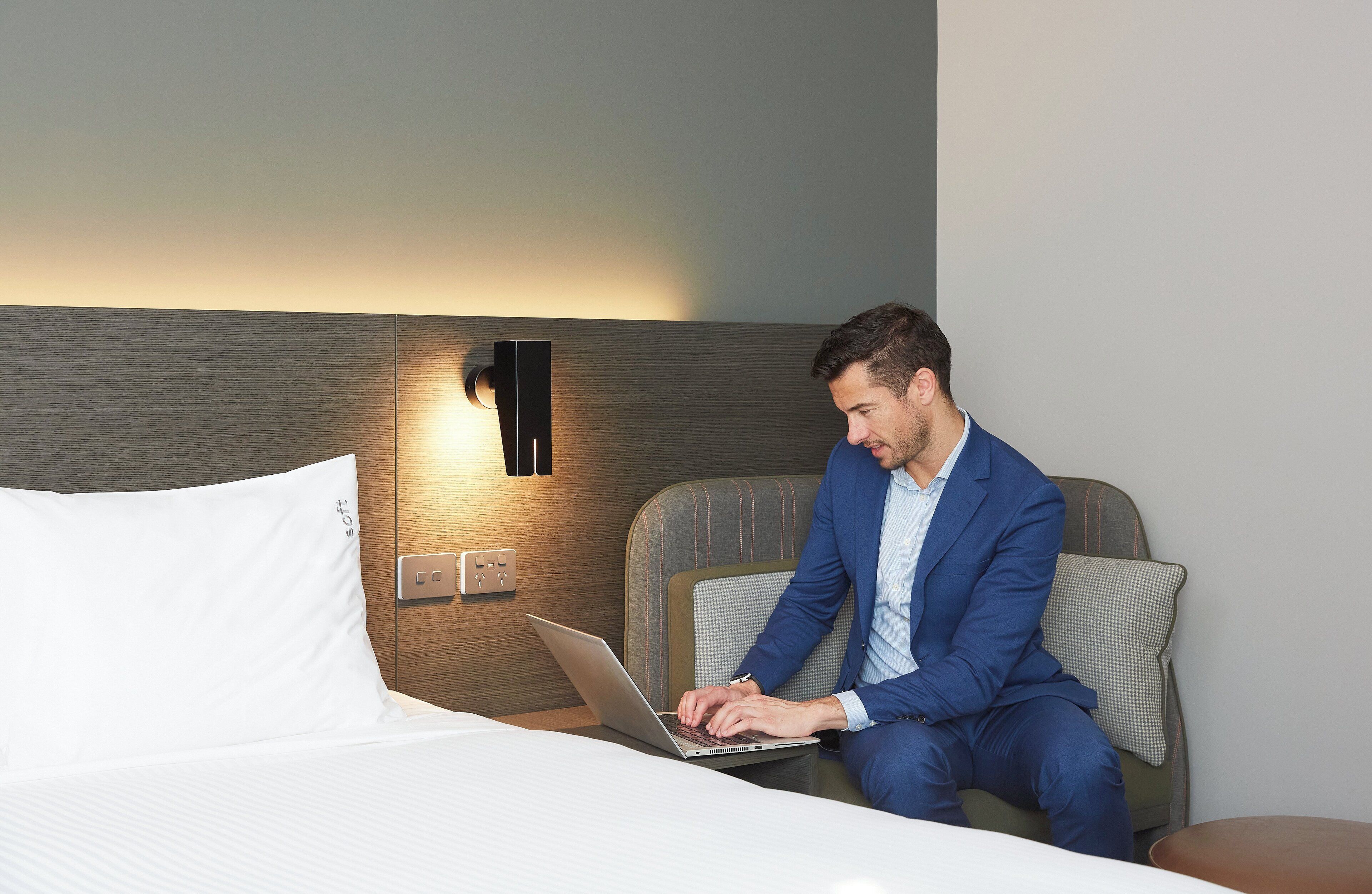 En mand i en blå jakkesæt bruger en bærbar computer i en hotelseng med et træhovedgærde og en lampe monteret på væggen.