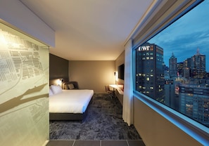 A hotel room with a bed, a desk, a chair, a large window with a city view, and a patterned wall. - Holiday Inn Express Melbourne Southbank by IHG (Southbank)