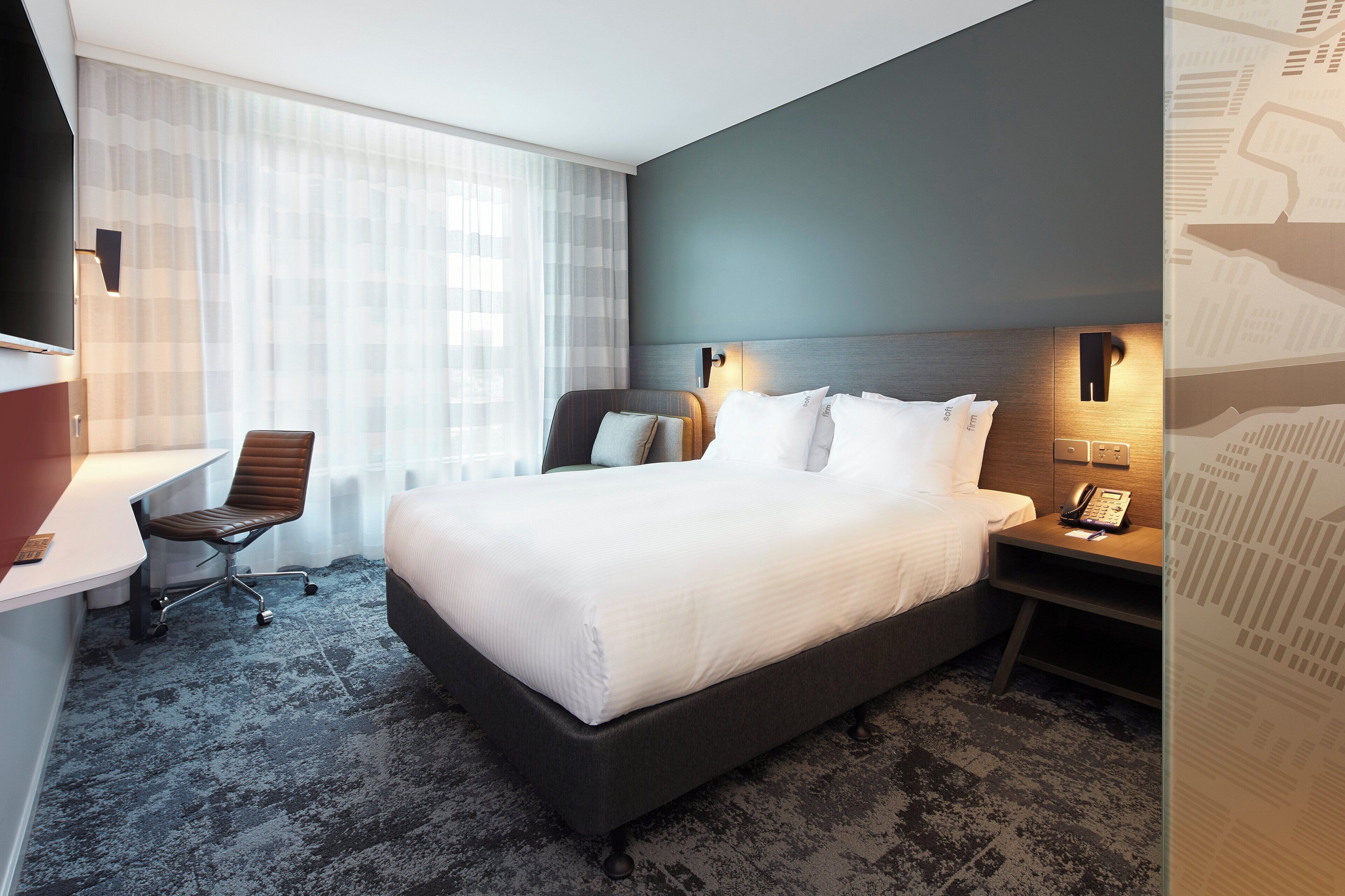 A hotel room with a large bed, a desk with a chair, a bedside table with a phone, and a window with sheer curtains.