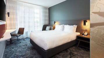 A hotel room with a large bed, a desk with a chair, a bedside table with a phone, and a window with sheer curtains.