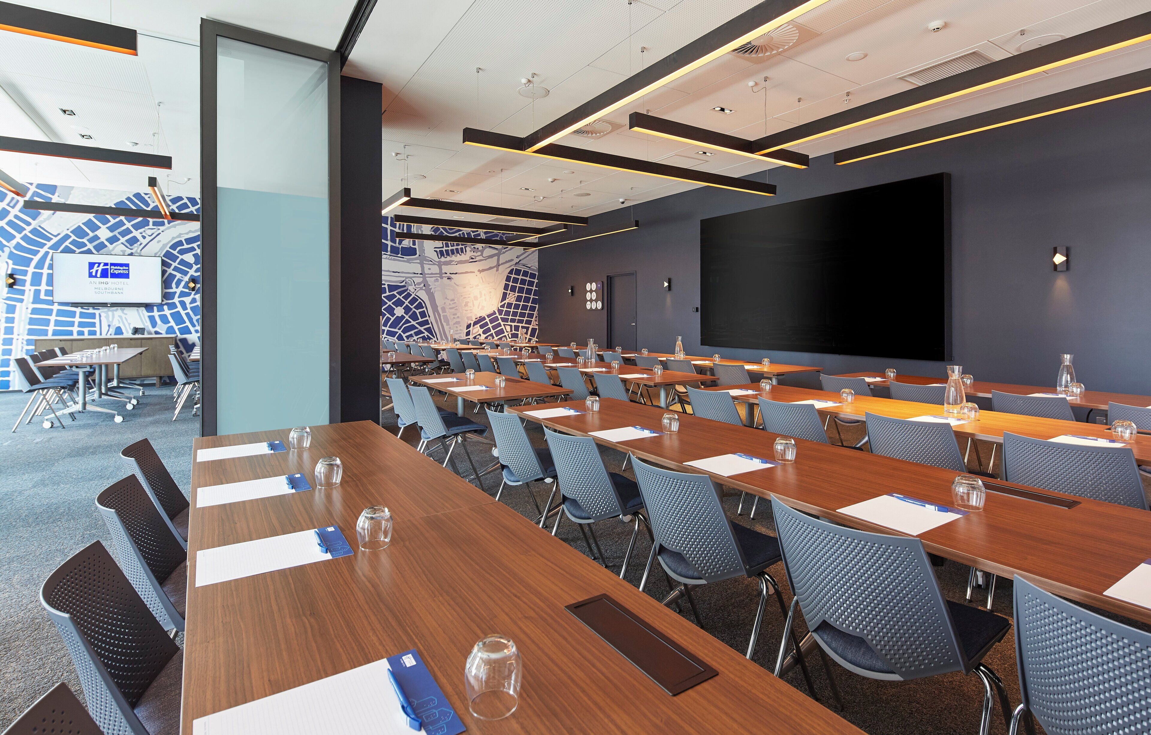 A conference room with multiple tables and chairs, a large screen, and a patterned wall.