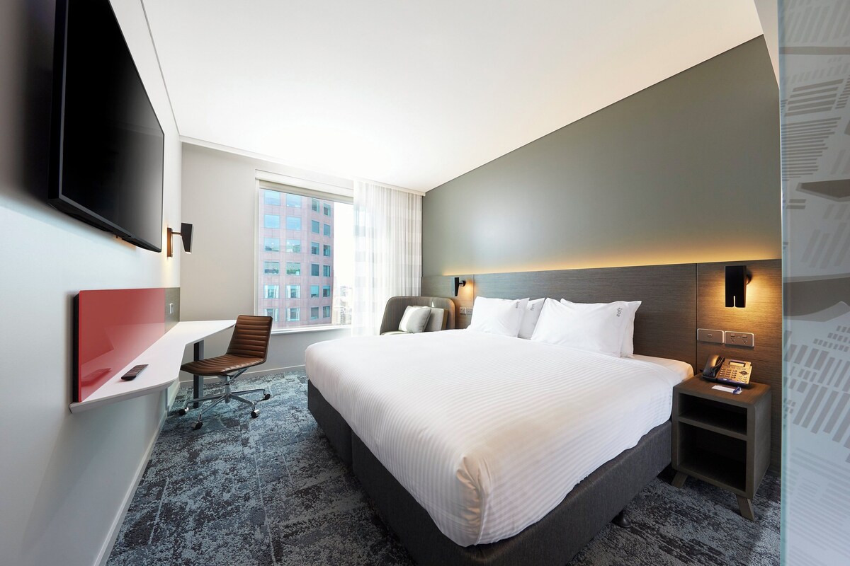A hotel room with a bed, a desk, a chair, a TV, a nightstand, and a window.