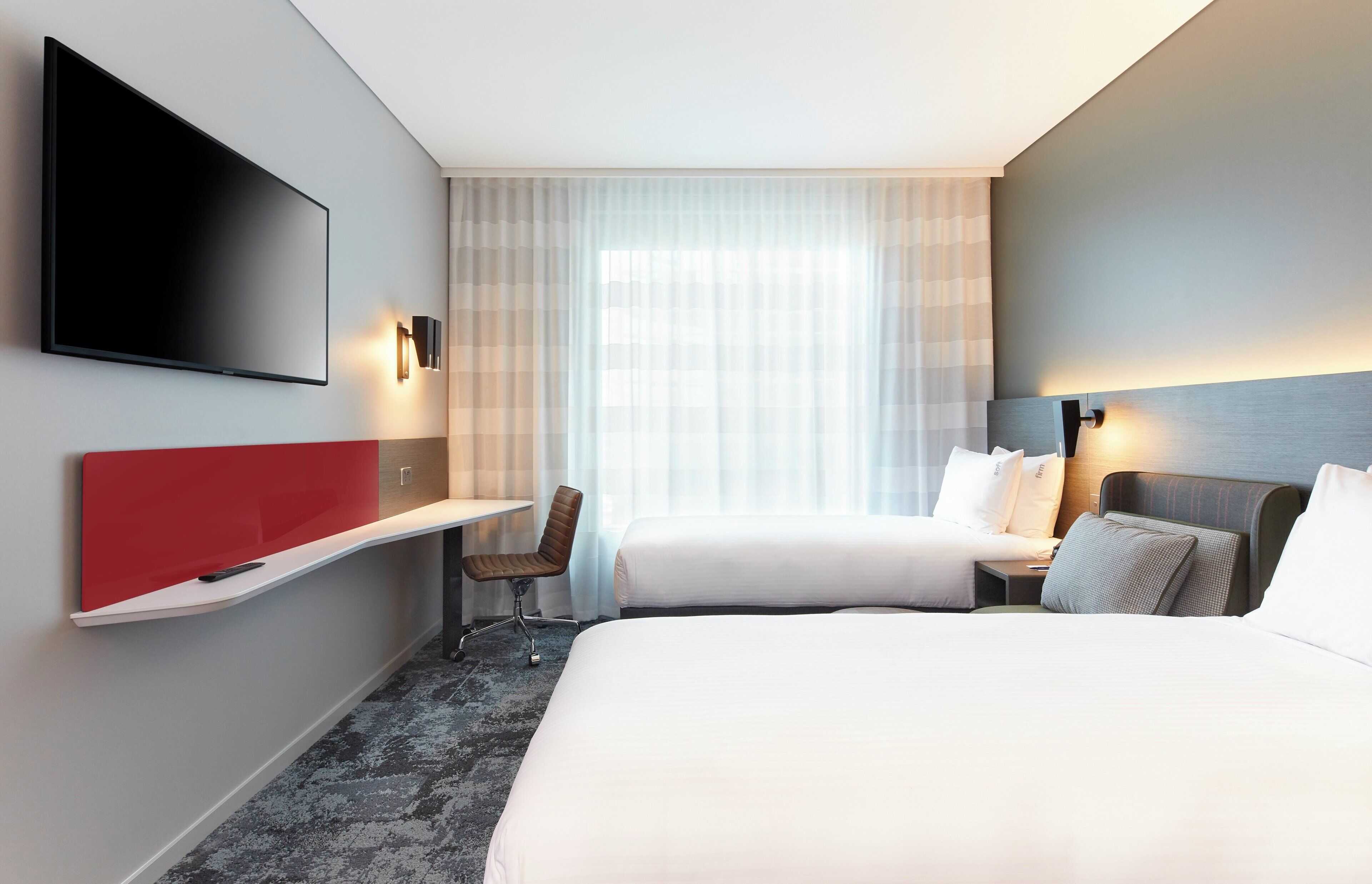 A hotel room with a flat-screen TV, a desk, two beds, and a large window with sheer curtains.