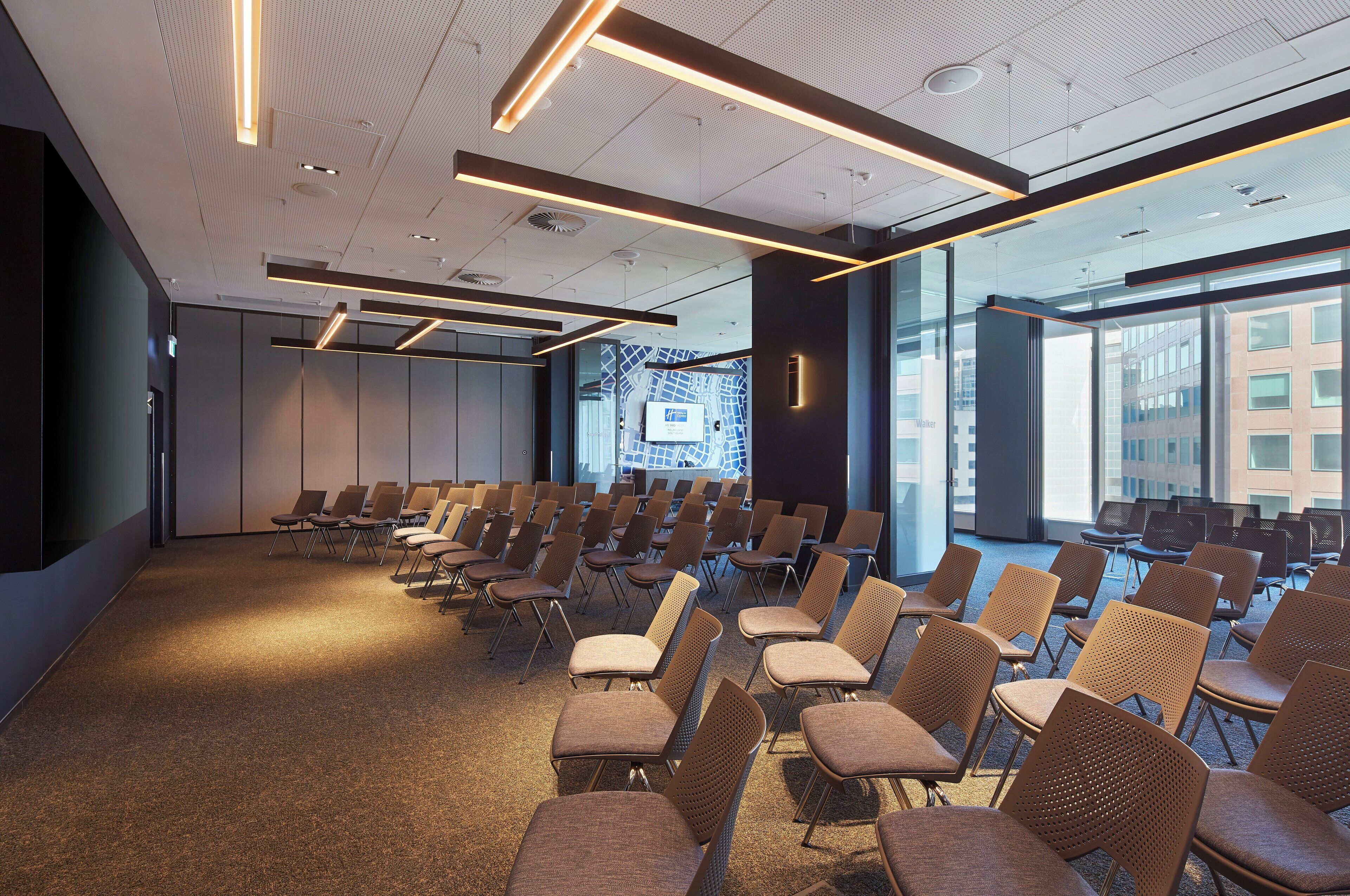 A conference room with rows of chairs, a large window with a cityscape view, and modern ceiling lighting.