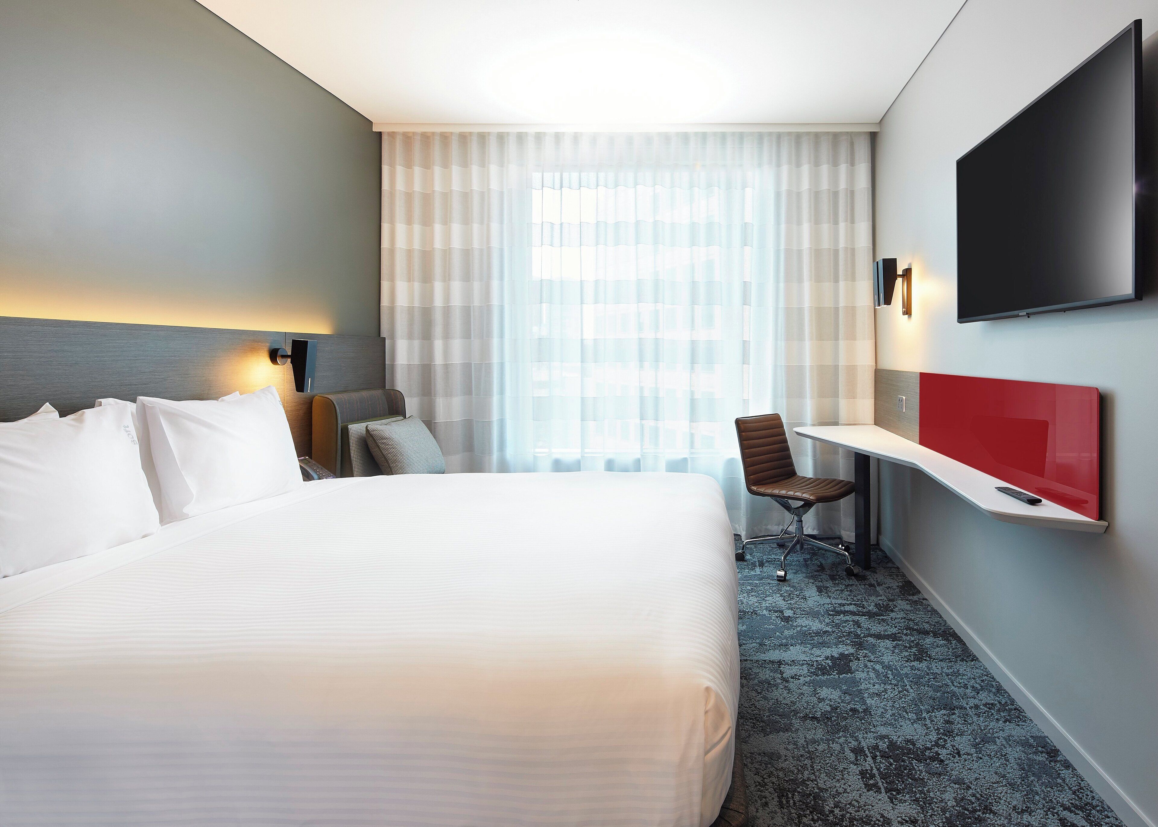 A hotel room with a large bed, a desk with a chair, a TV mounted on the wall, and a window with sheer curtains.