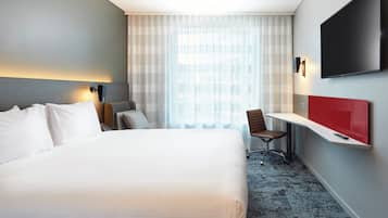 A hotel room with a large bed, a desk with a chair, a TV mounted on the wall, and a window with sheer curtains.