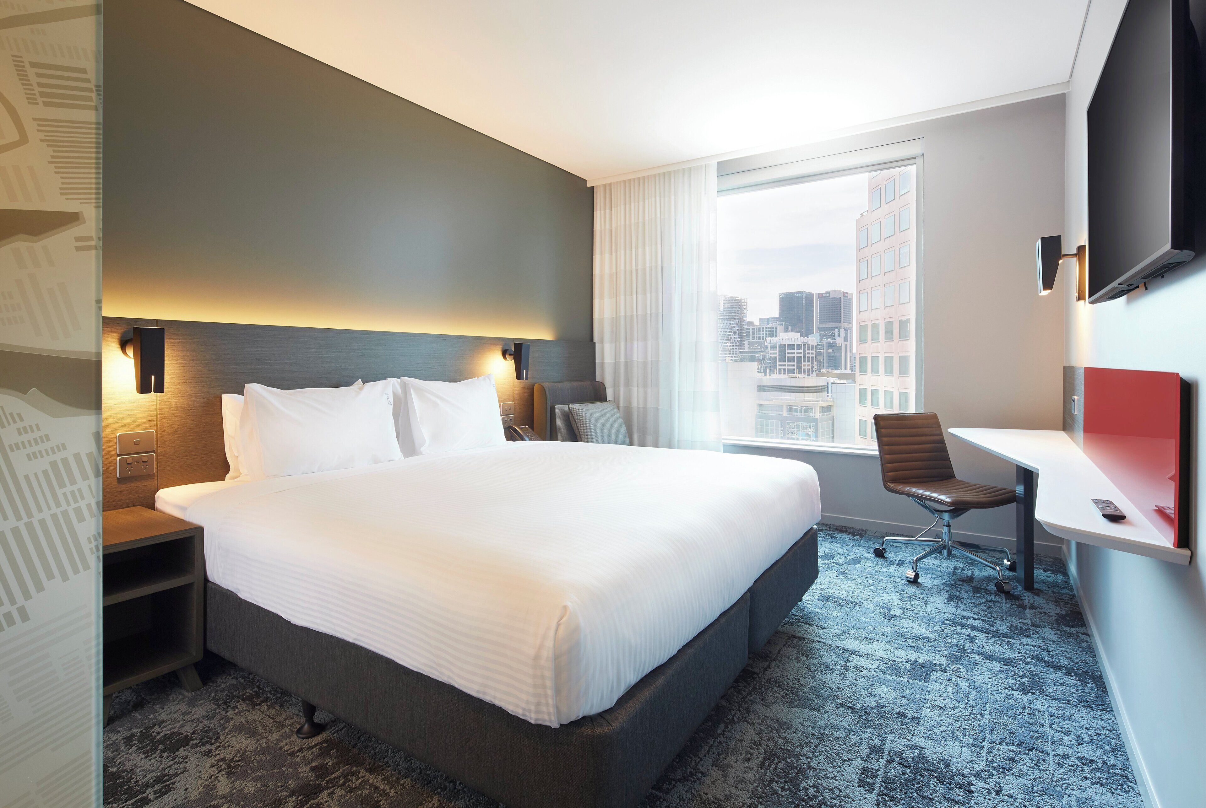 A hotel room with a large bed, a desk, a chair, a TV, and a window with a city view.