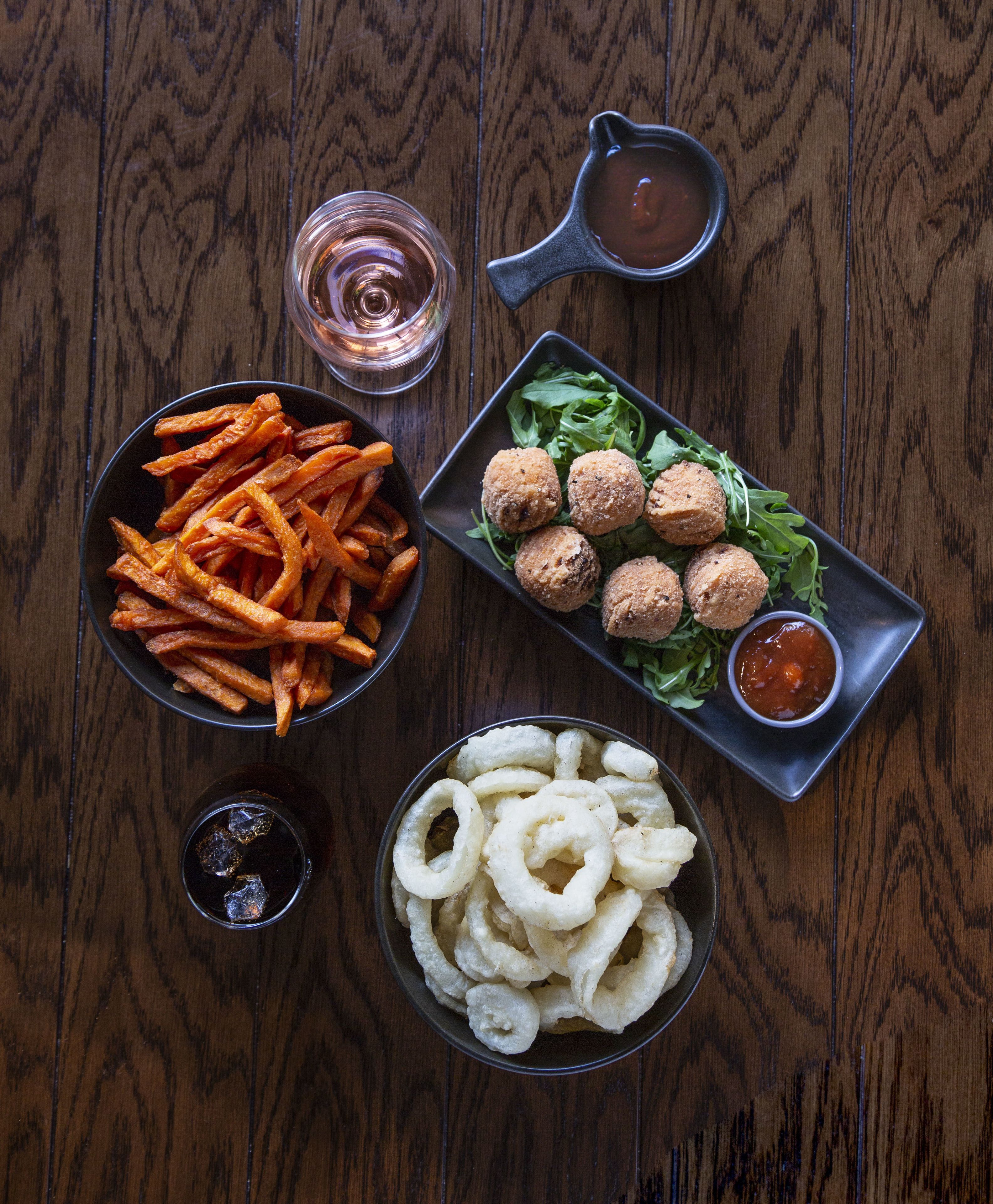 Una mesa de madera con cuencos de papas fritas, croquetas, aros de cebolla y un tazón pequeño de salsa.