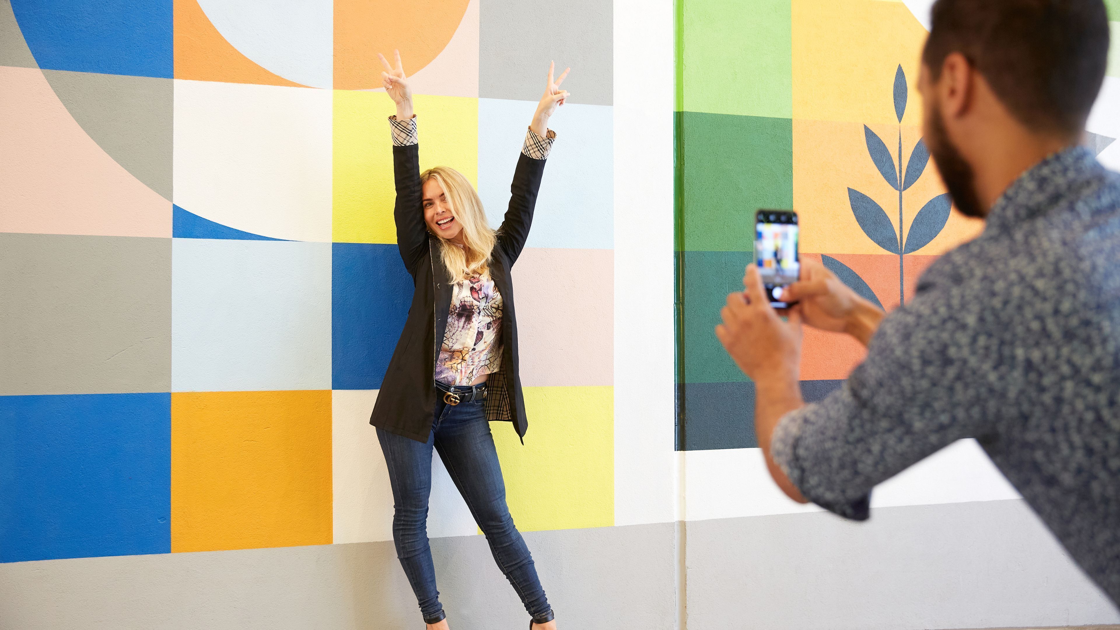 Una persona fotografiando a otra frente a una pared con diseños geométricos y colores vibrantes.