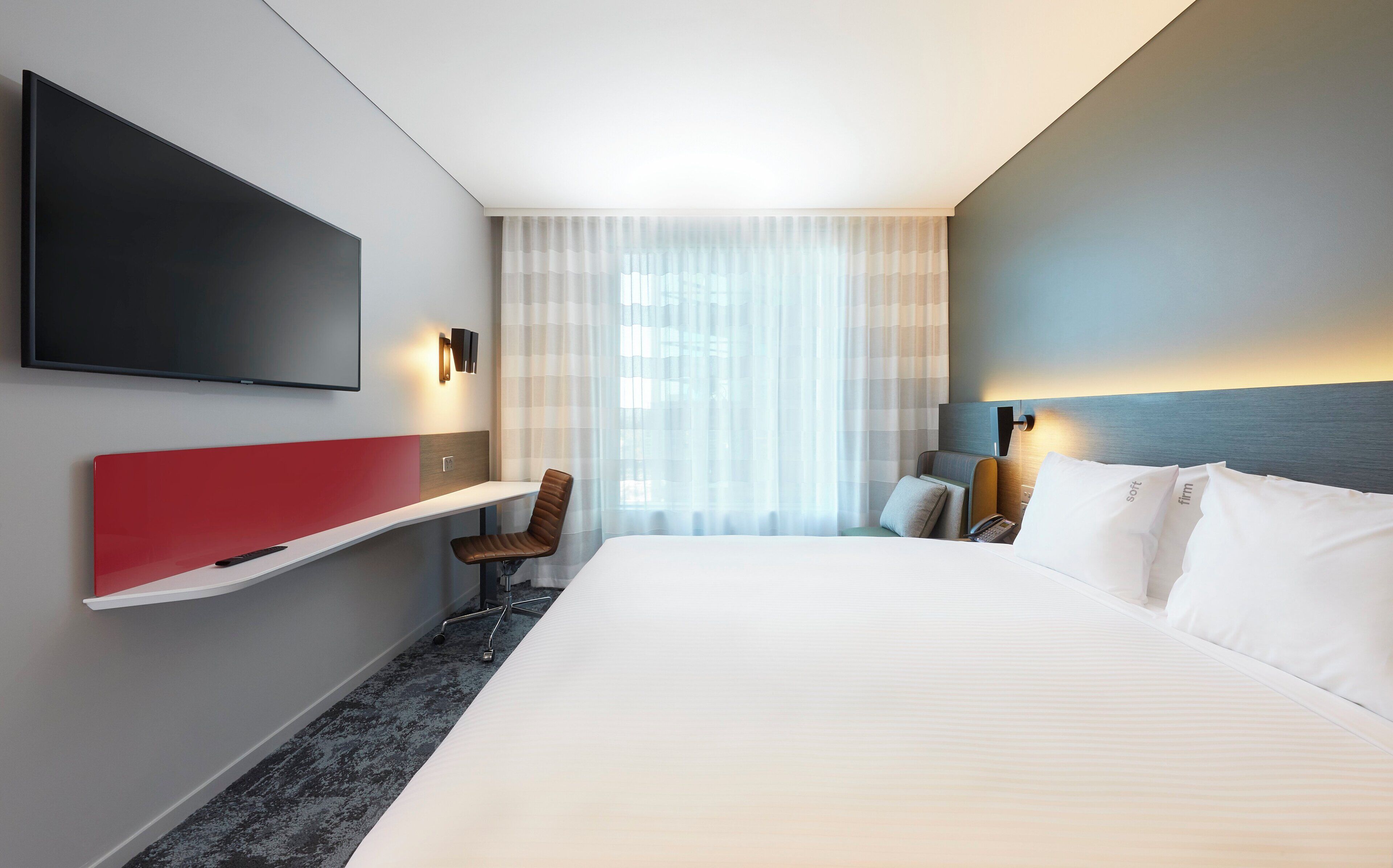 Habitación de hotel con una cama grande, televisor montado en la pared, escritorio con silla y ventana con cortinas translúcidas.