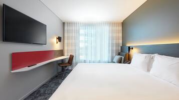 Habitación de hotel con una cama grande, televisor montado en la pared, escritorio con silla y ventana con cortinas translúcidas.