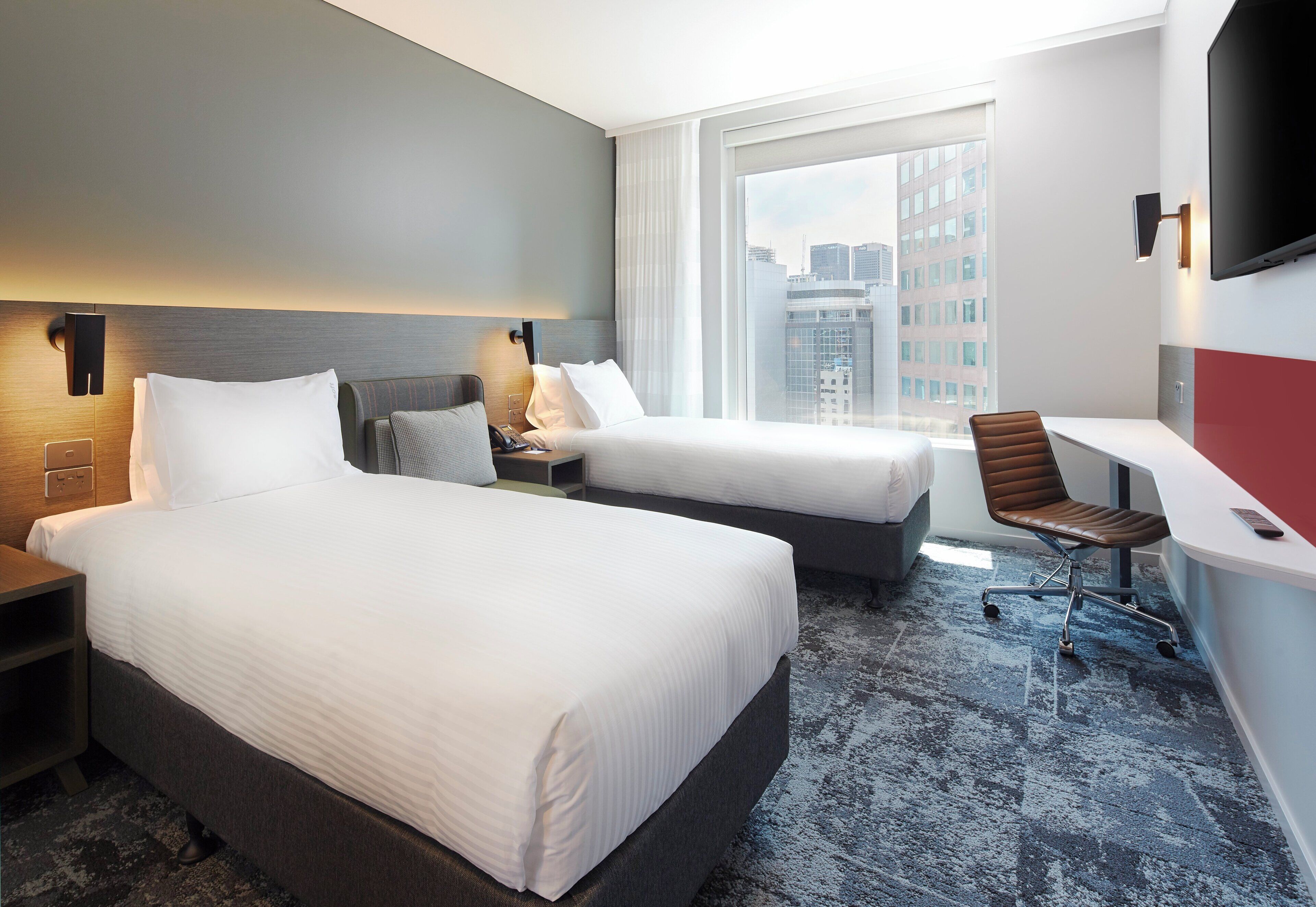 A hotel room with two beds, a desk, a chair, and a large window with a city view.