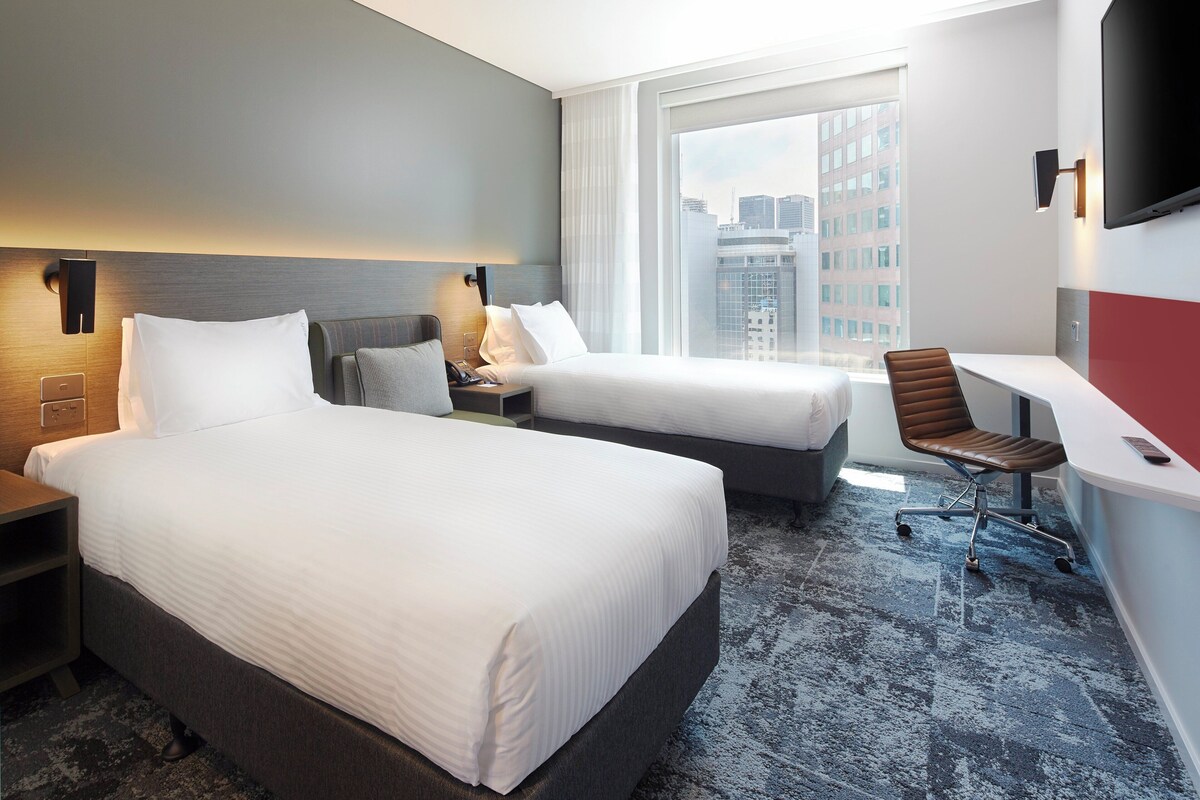 A hotel room with two beds, a desk, a chair, and a large window with a city view.