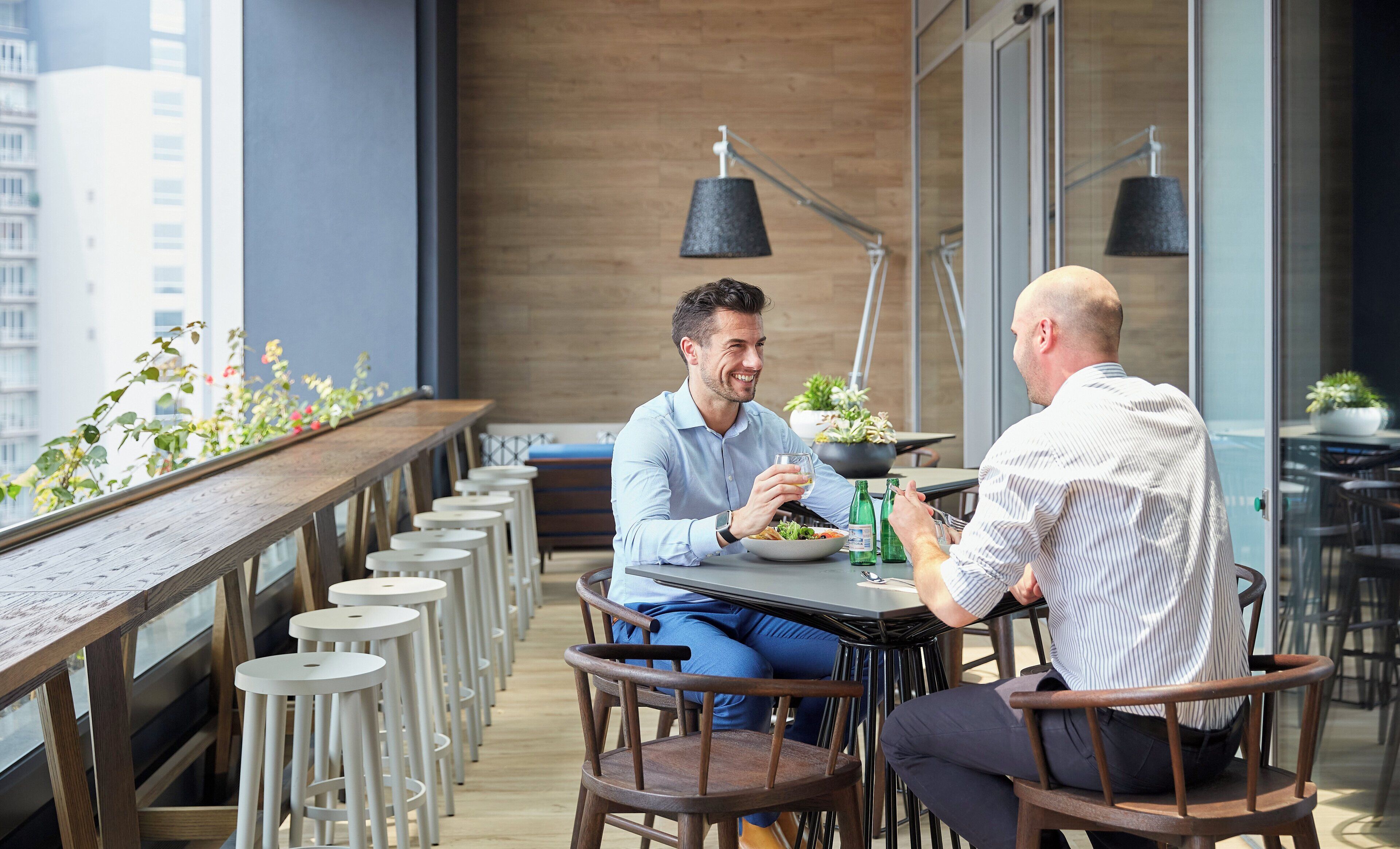 Two people seated at a table with a view of the city, wooden chairs, and potted plants.