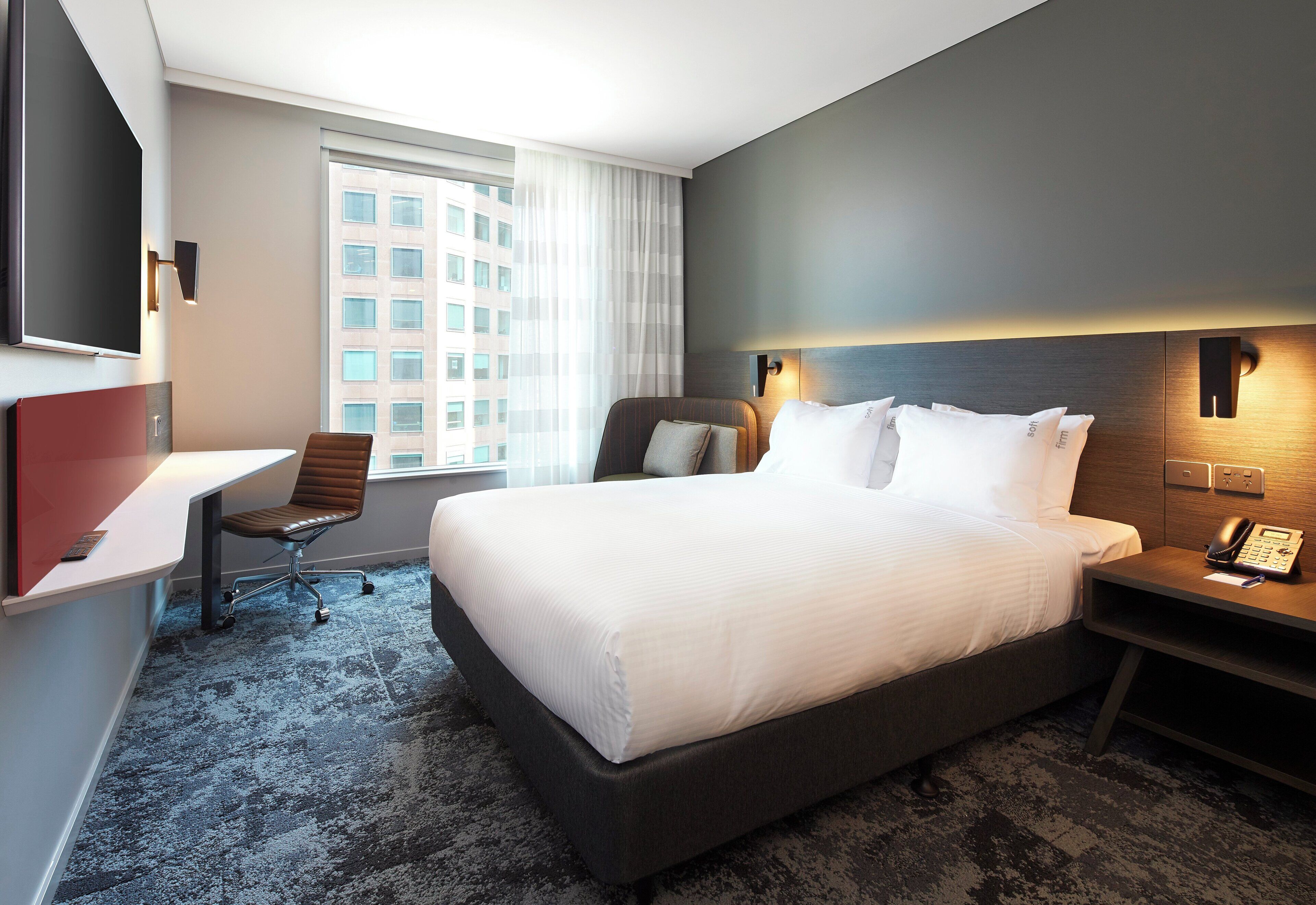 A hotel room with a large bed, a desk, a chair, a TV, and a window with a view of buildings.