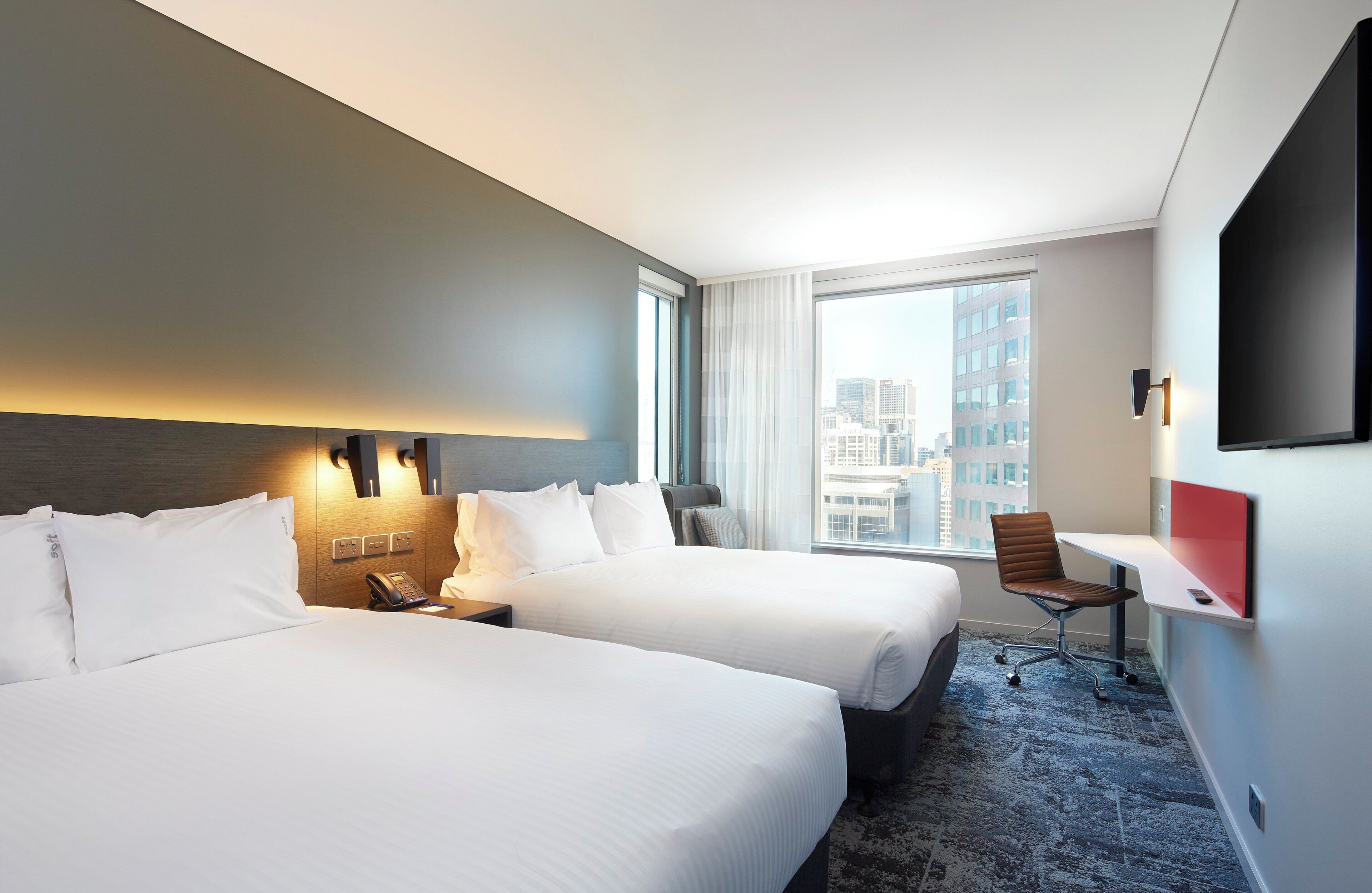 A hotel room with two beds, a desk, a chair, a phone, and a large window with a city view.