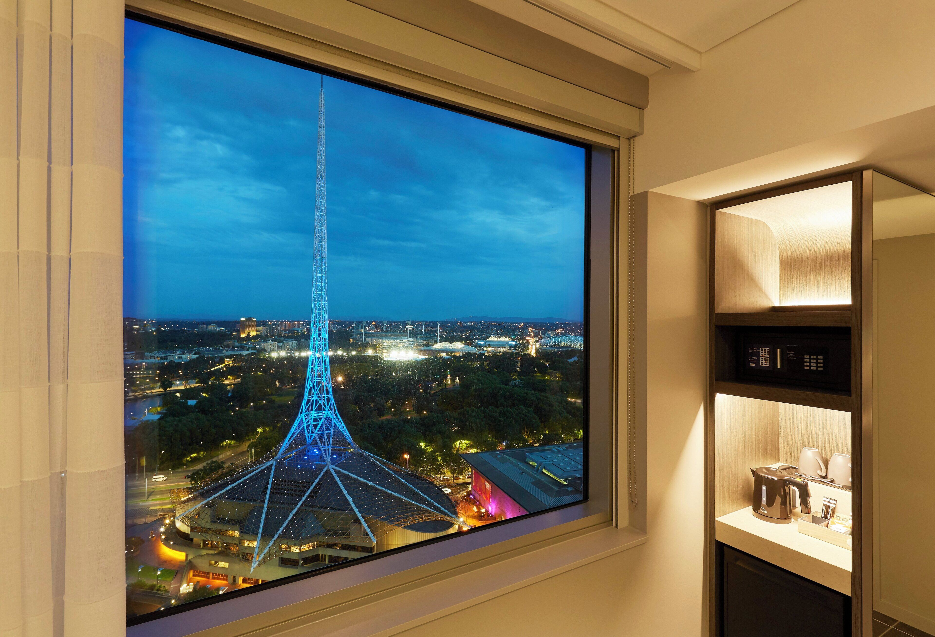 Quarto de hotel com uma grande janela que oferece vista para a paisagem urbana à noite, destacando uma imponente estrutura em torre.