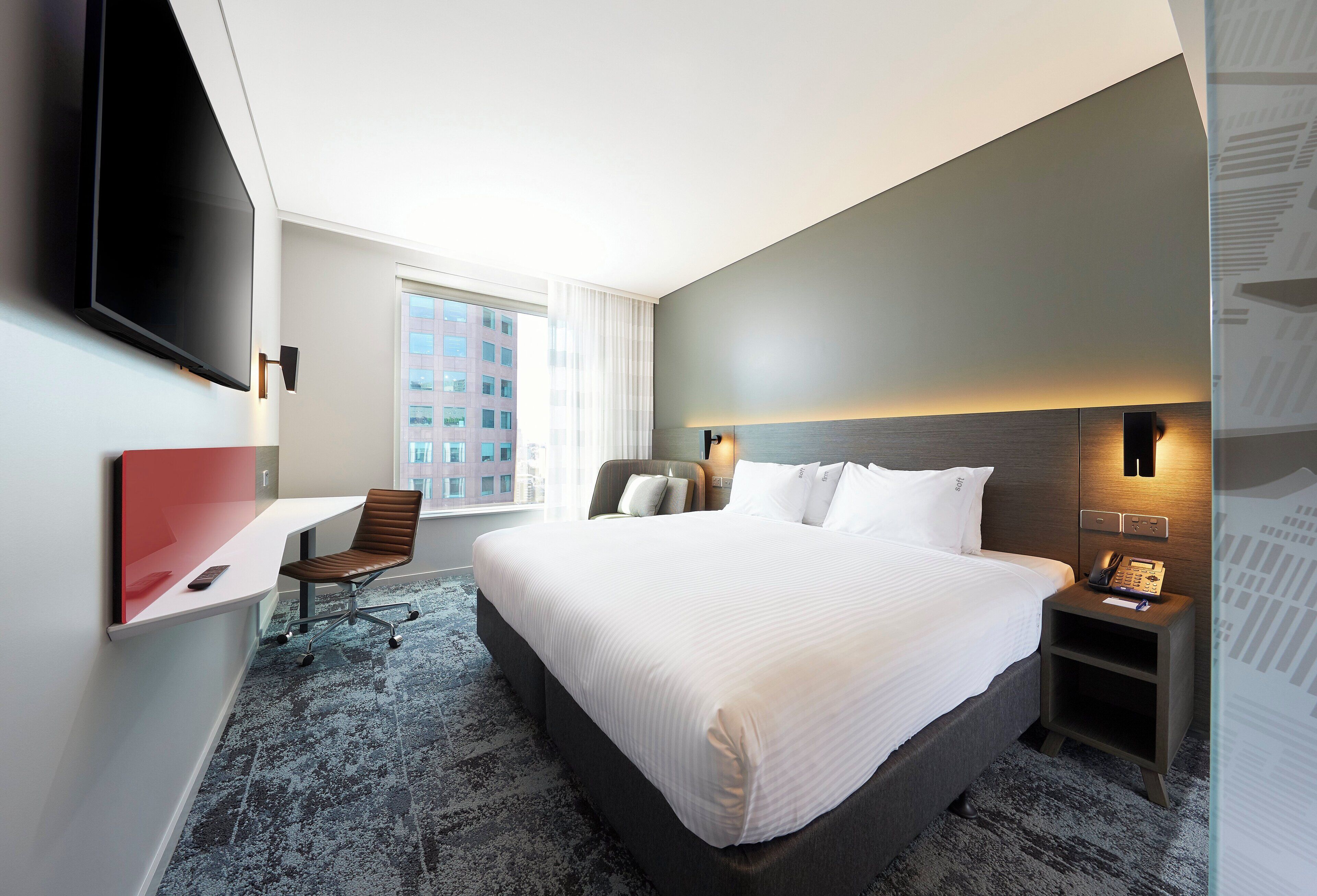 Une chambre d’hôtel avec un grand lit, un bureau avec une chaise, un téléviseur à écran plat fixé au mur, une table de chevet avec un téléphone et une fenêtre avec des rideaux.
