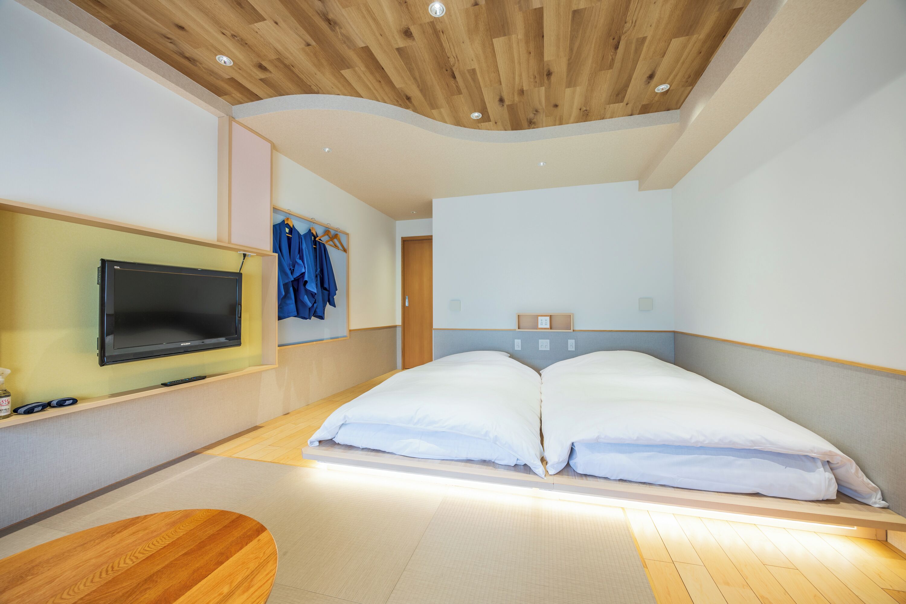 Una habitación de hotel moderna con techo de madera, un televisor de pantalla plana montado en la pared, una cama con ropa blanca y suelo de madera.