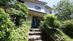 Un bâtiment à deux étages avec une porte bleue et une enseigne au-dessus. Des marches en pierre mènent à l'entrée, qui est entourée de verdure.