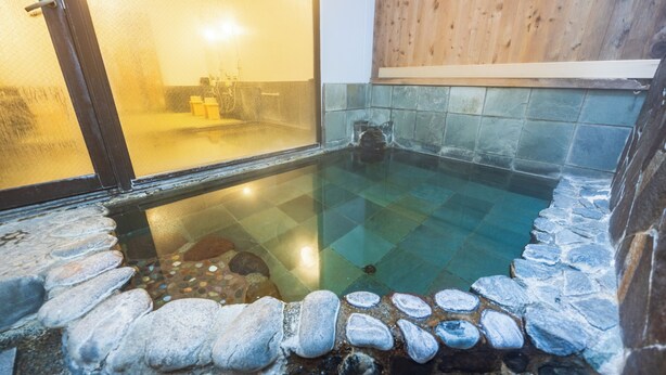 Un bain thermal, alimenté par une source d’eau chaude, avec des marches en pierre menant à l’eau, entouré de murs en bois et doté d’un sol carrelé.
