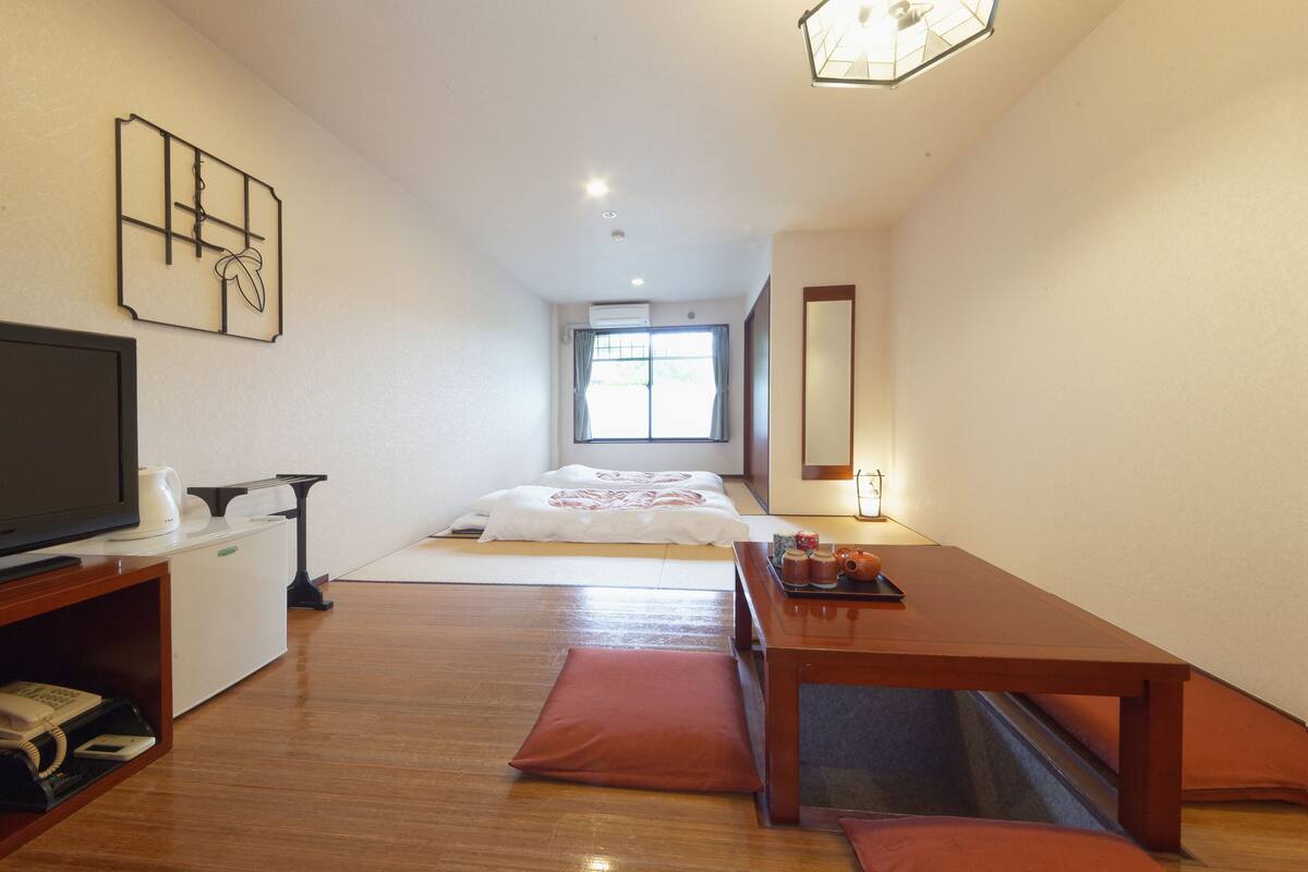 Une chambre d’hôtel de taille réduite, comprenant un lit, une télévision, une petite table avec un plateau contenant divers objets, un meuble en bois et une fenêtre avec des rideaux.