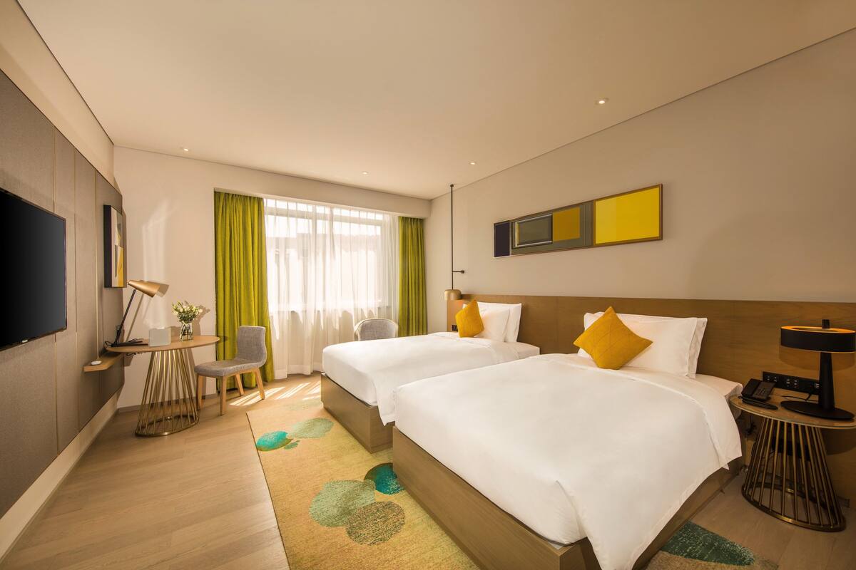 A hotel room with two beds, a desk, a chair, a TV, and a window with curtains.