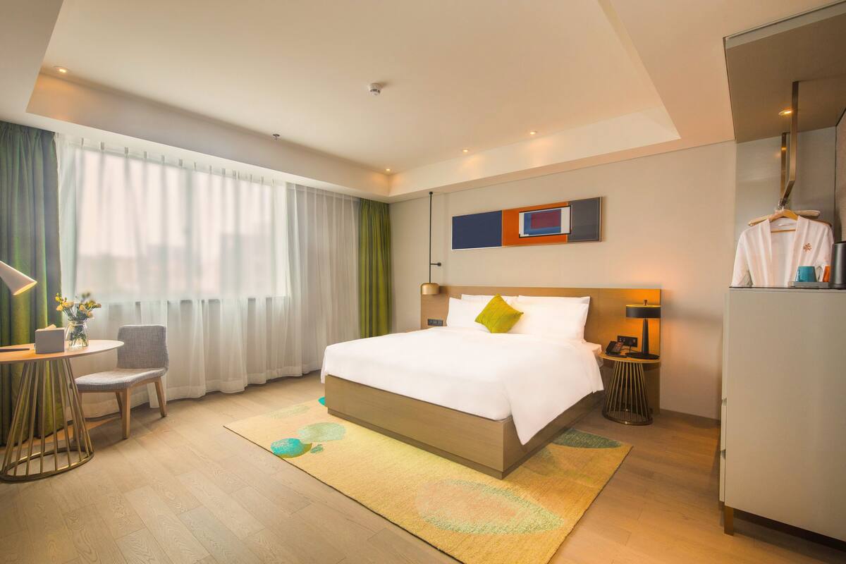 A hotel room with a bed, a desk with a chair, a wardrobe, and a window with curtains.