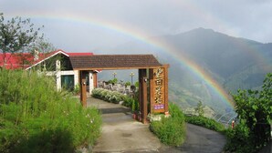 Um arco de madeira com uma placa, que leva a um edifício com telhado vermelho, cercado por vegetação e uma montanha ao fundo.