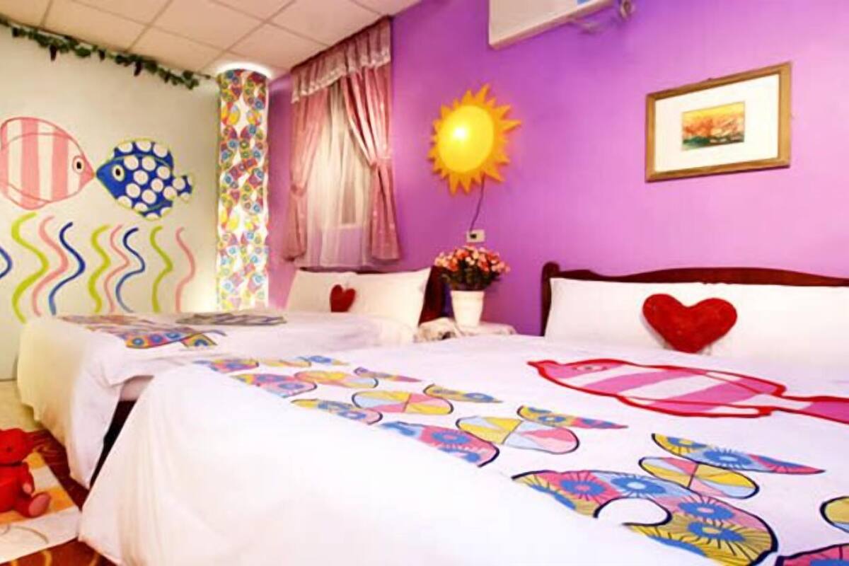 Une chambre avec deux lits, des œuvres d’art colorées accrochées au mur, une lampe de luminothérapie et des coussins en forme de cœur.