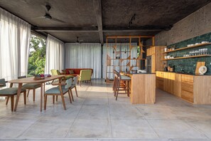 A spacious kitchen with wooden cabinets, a dining area with a table and chairs, and a view of greenery outside. - Balian Surf Villas (Selemadeg)