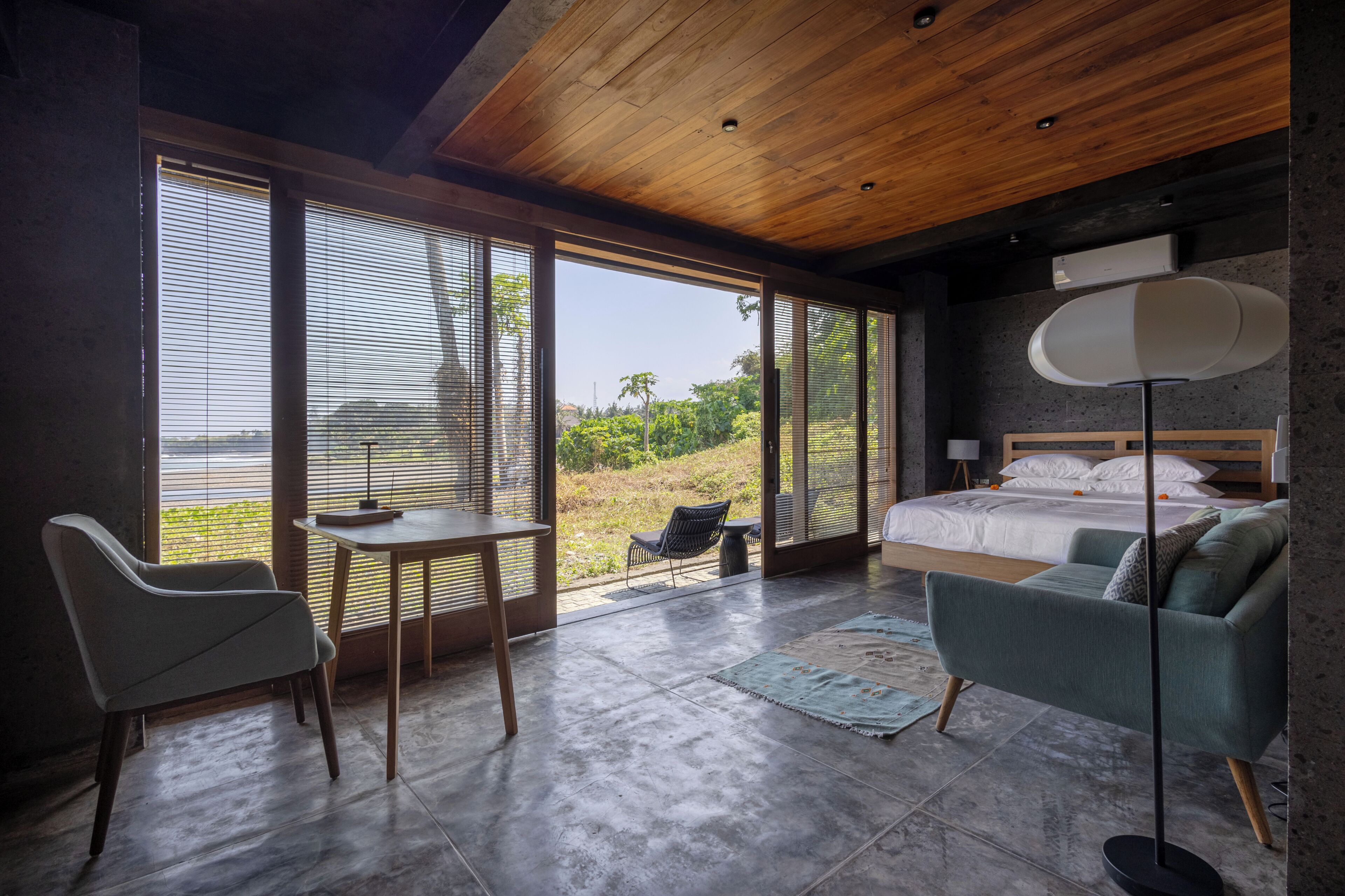 A modern bedroom with a large bed, a sofa, a table, and a chair. There is a view of the outdoors through large windows.