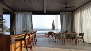 A modern kitchen with a bar area, large windows with curtains, and a view of the ocean. - Balian Surf Villas (Selemadeg)