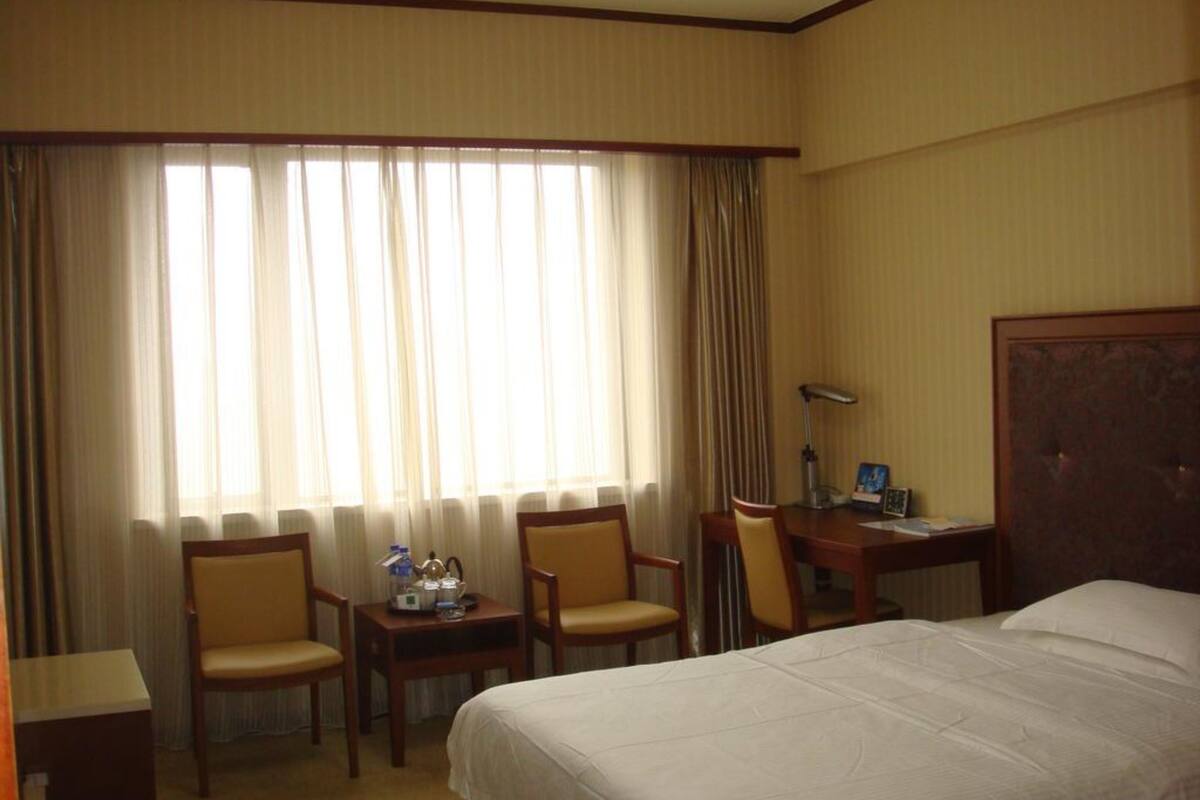 Ein Hotelzimmer mit einem Bett, zwei Stühlen, einem Schreibtisch, einer Lampe und einem Fenster mit Vorhängen.