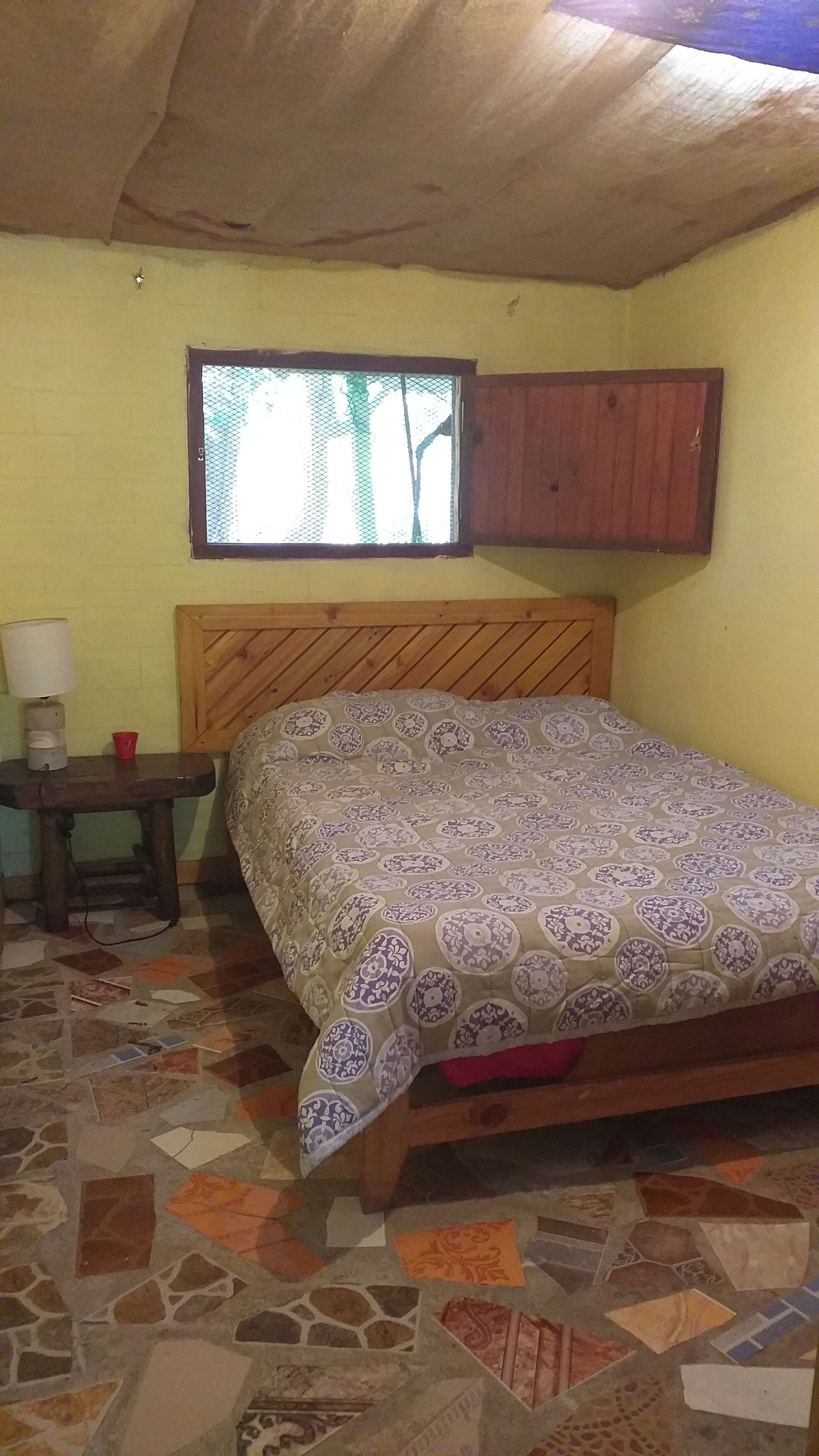 A bedroom with a wooden bed, a nightstand with a lamp, a window with a wooden frame, and a patterned floor.