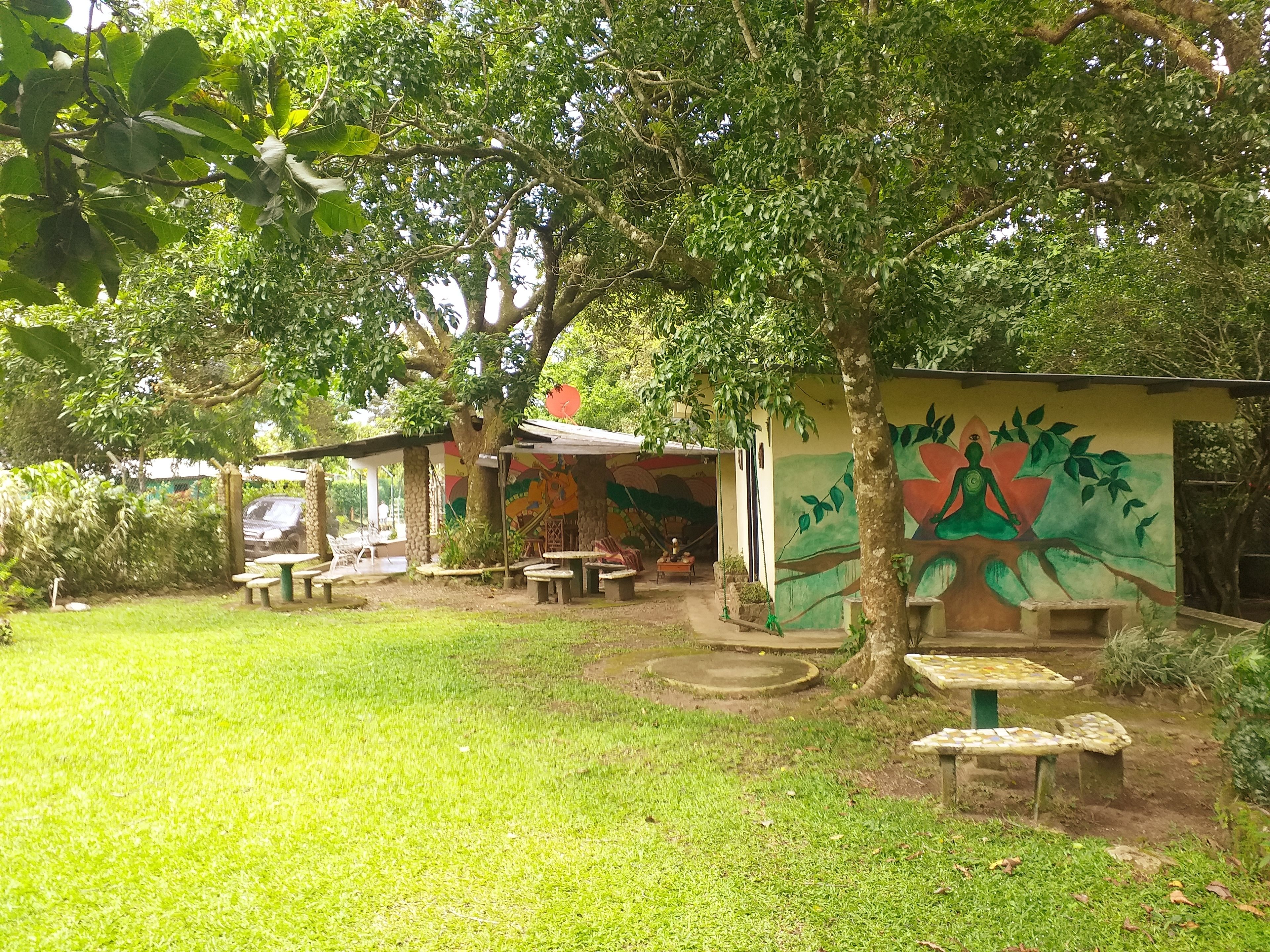 Un espacio exterior tranquilo con césped, un mural y un pequeño pabellón con mesas y sillas.