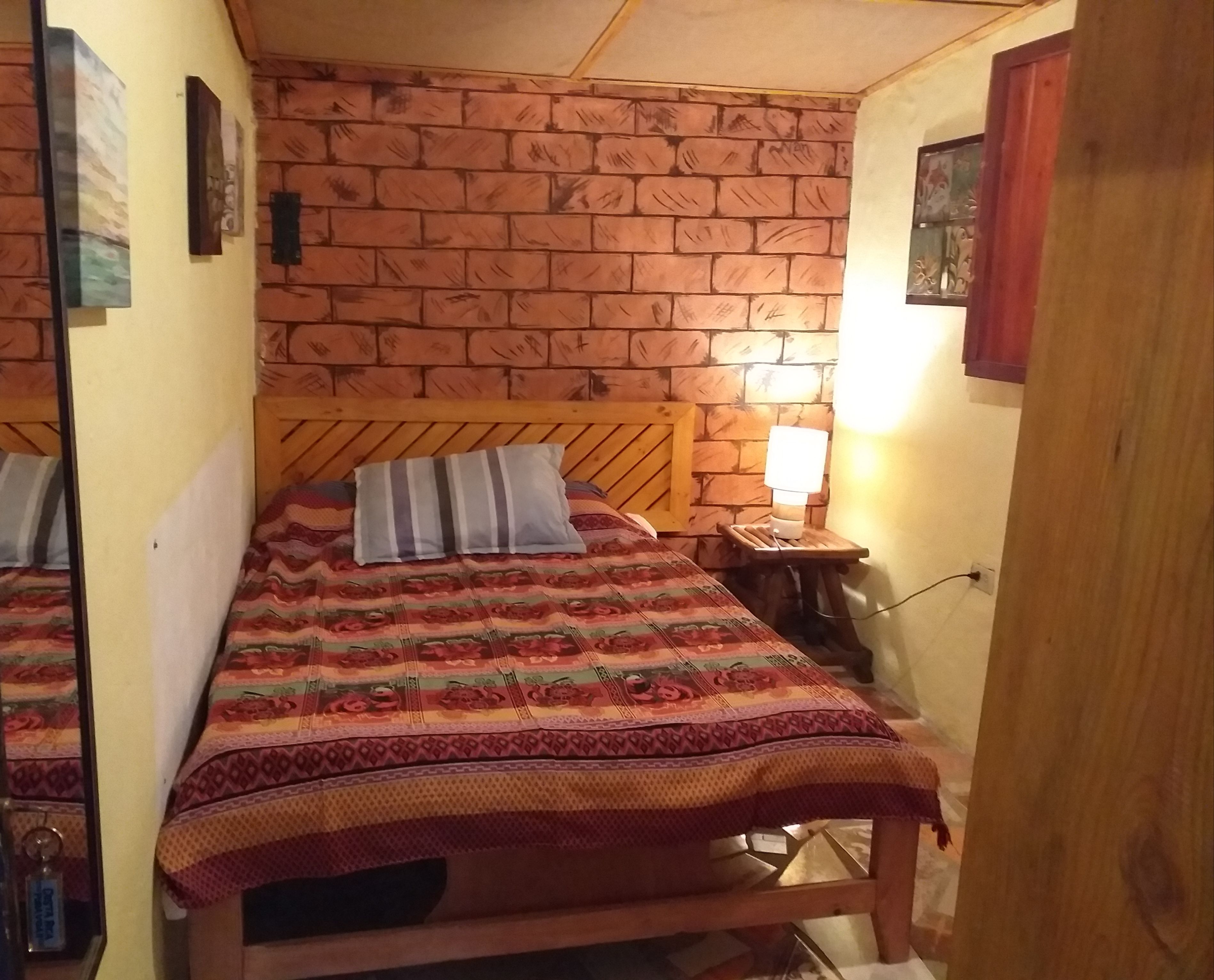 A room with a brick-patterned wall, a bed with a floral bedspread, a bedside lamp, and a small wooden nightstand.