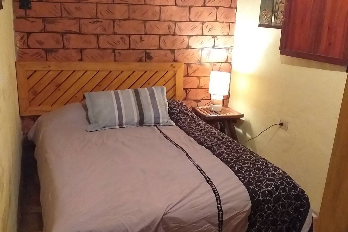 Ett litet, ombonat rum med en vägg som har ett tegelstensmönster, en sänggavel i trä, en säng med en randig kudde, ett mönstrat överkast och en sänglampa.