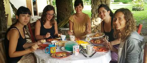 A group of people sitting around a table with various dishes and drinks, in an outdoor setting with trees and a stone wall in the background.