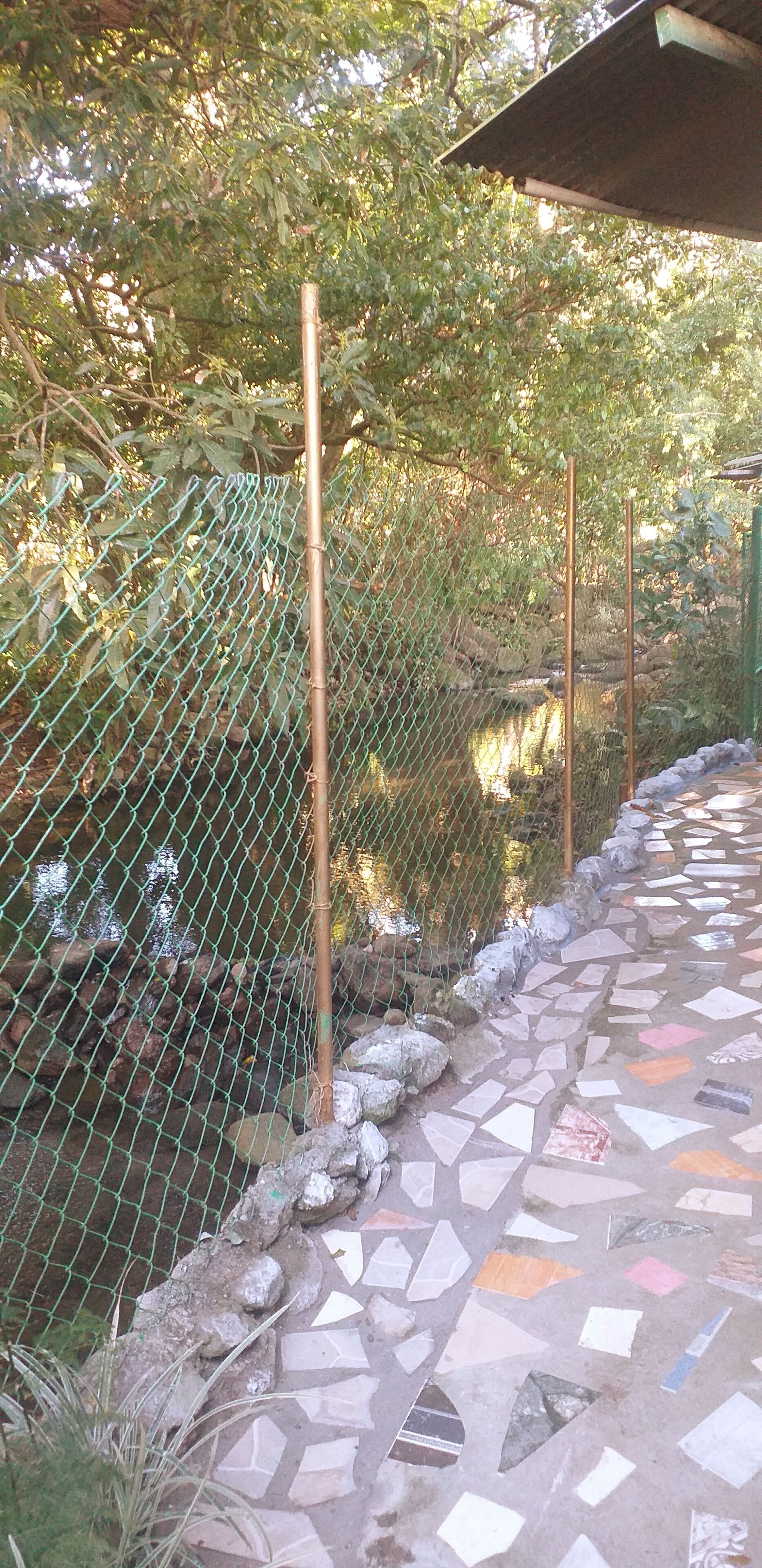 Un percorso lastricato con mosaici corre lungo un recinto in rete metallica con un palo di legno, diretto verso un corpo d'acqua.