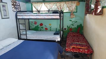 Una habitación con dos literas, una con un colchón estampado con motivos florales y la otra con un colchón liso. Hay una cama individual con una colcha azul. Las paredes están pintadas de verde y hay una ventana con cortina.