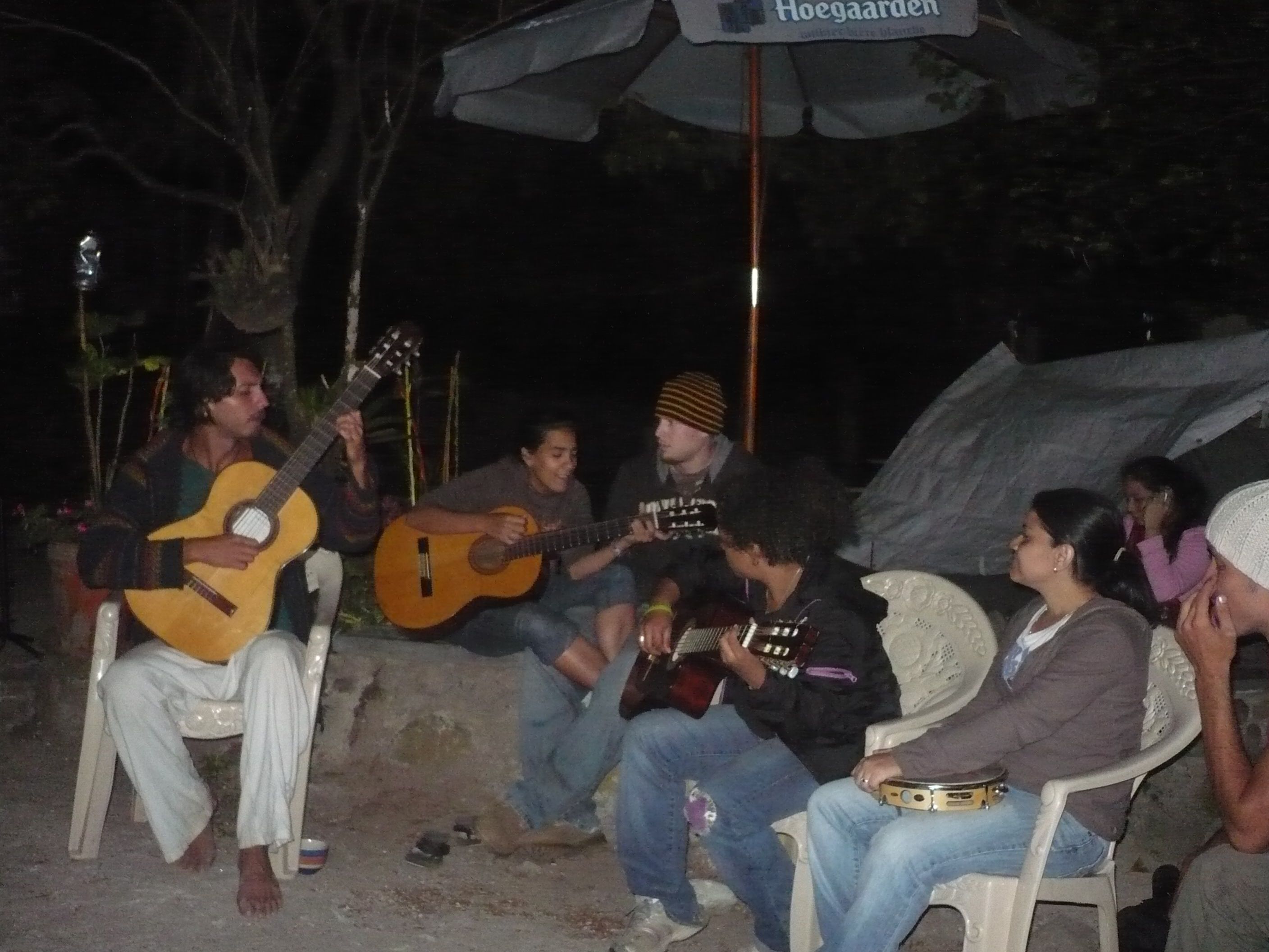 Un grupo de personas sentadas en sillas de plástico al aire libre por la noche, algunas tocando guitarras.